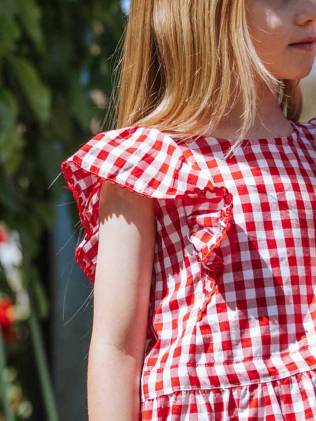 The child, dressed in a delightful Red Gingham Lace Edge Dress by Red Gingham, stands outdoors. The red and white checkered pattern adds a playful touch to the picturesque scene.