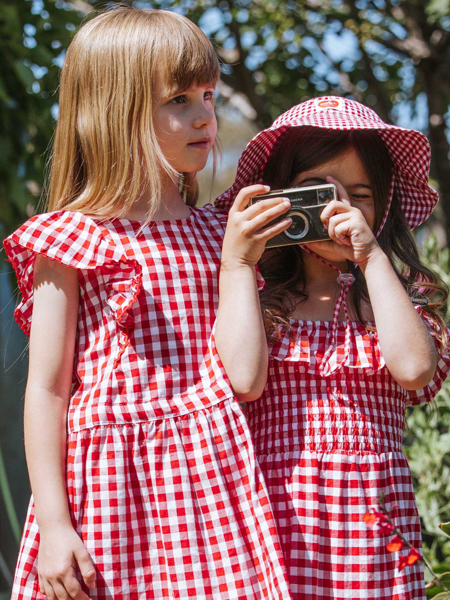 Two young girls in matching red checkered dresses made from 100% cotton stand outdoors among greenery. One is holding a camera to her eye, while the other adjusts her Red Gingham Bucket Hat, capturing their playful adventure in a hand-printed style.