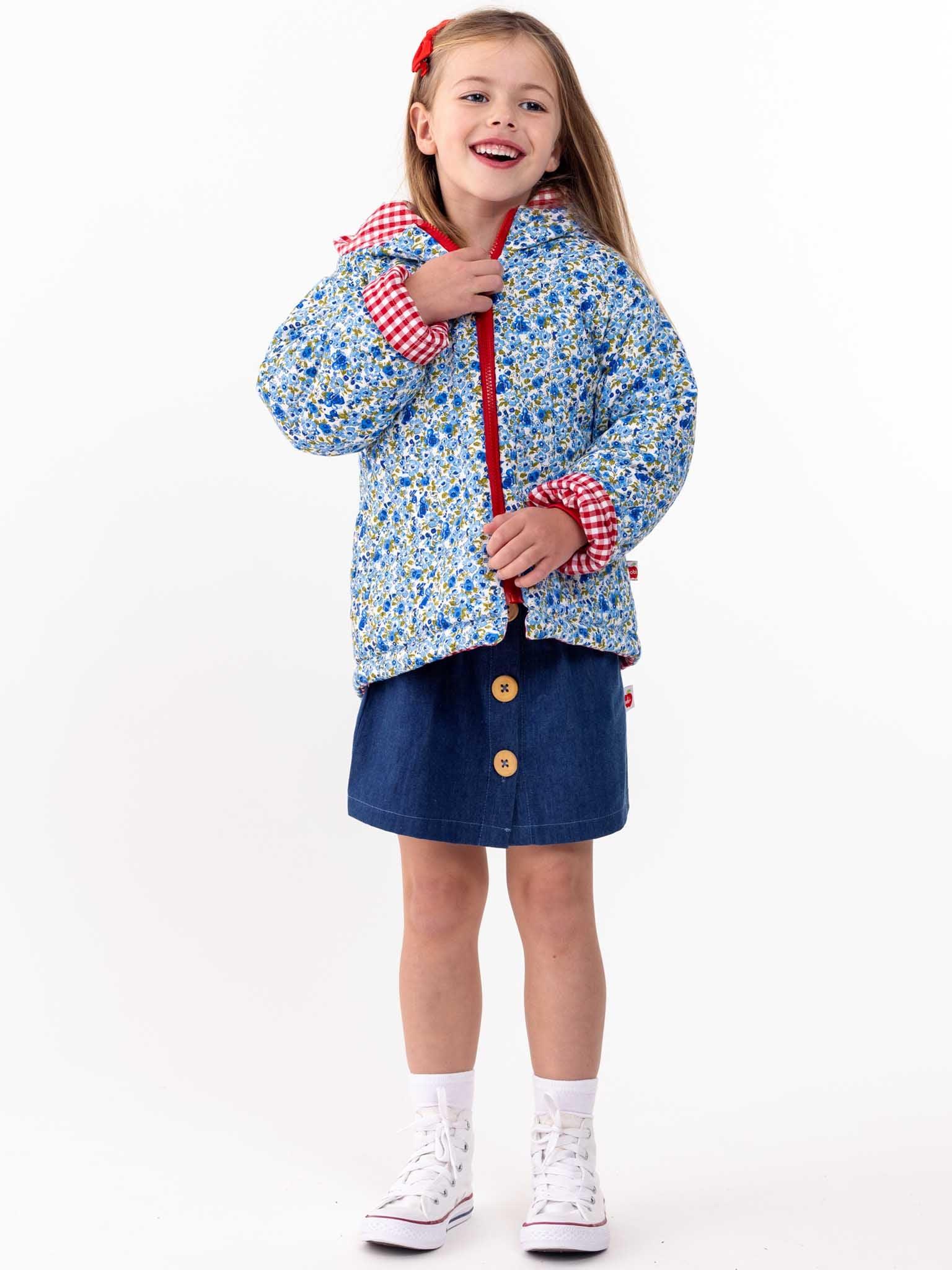 Young girl in an English Wildflowers floral quilted jacket and denim skirt smiling, holding an umbrella, standing against a white background.