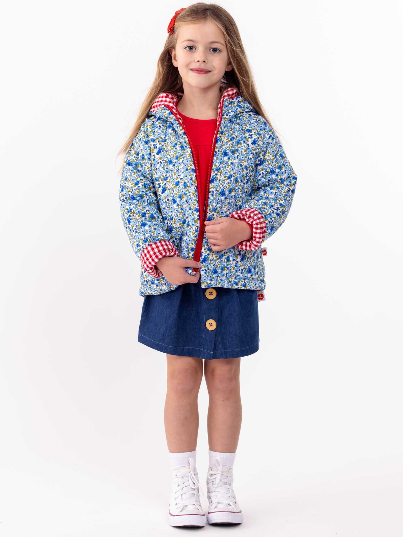 A young girl stands against a white background, wearing an English Wildflowers hooded reversible jacket, red shirt, denim skirt, and white sneakers. She has long brown hair and a red hairband.