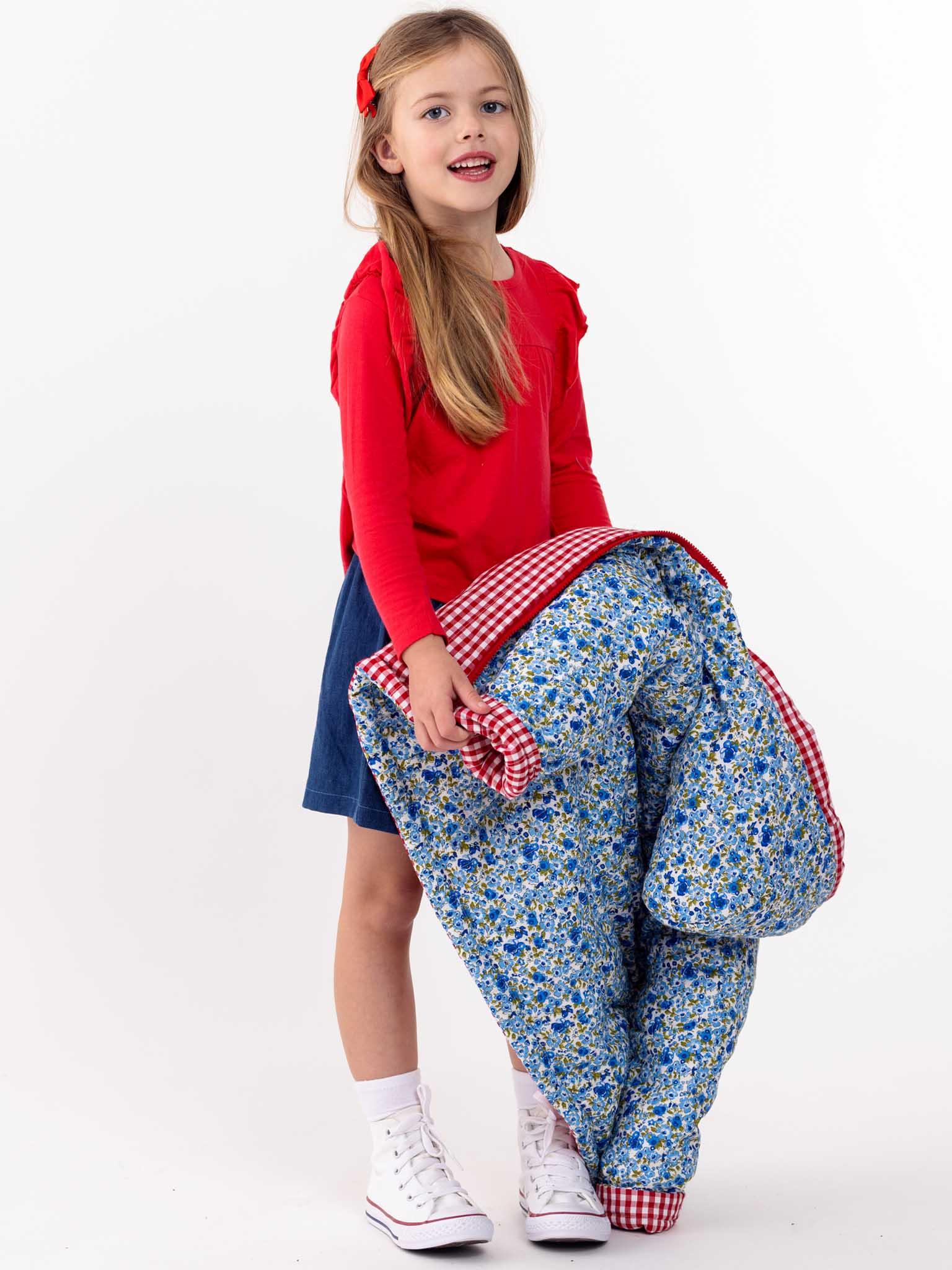 A young girl in a red shirt and blue skirt, holding an English Wildflowers English Flowers Hooded Reversible Jacket, stands against a white background, smiling.