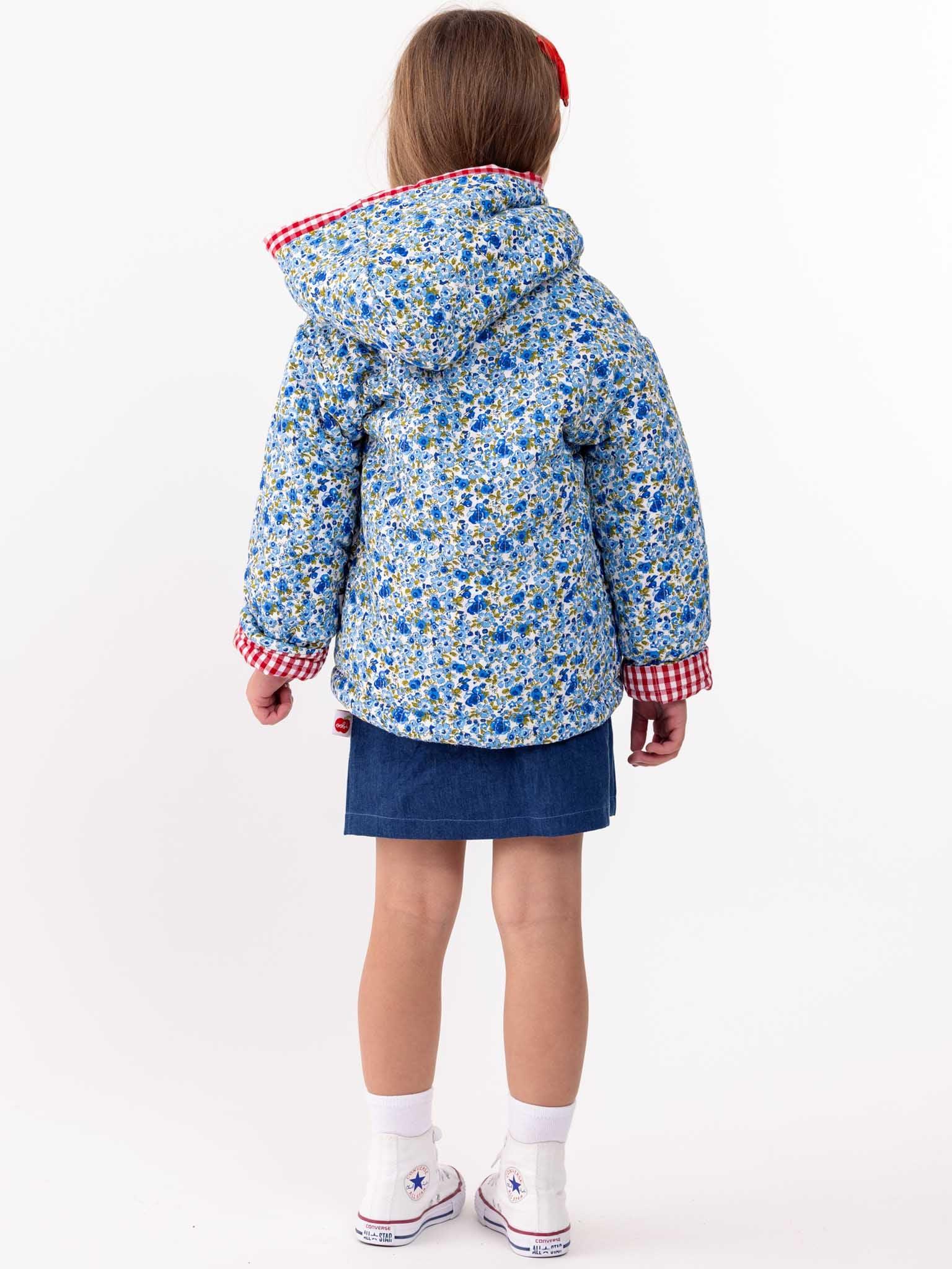 Young girl standing with her back to the camera, wearing a English Wildflowers' English Flowers Hooded Reversible Jacket and a blue skirt against a white background.