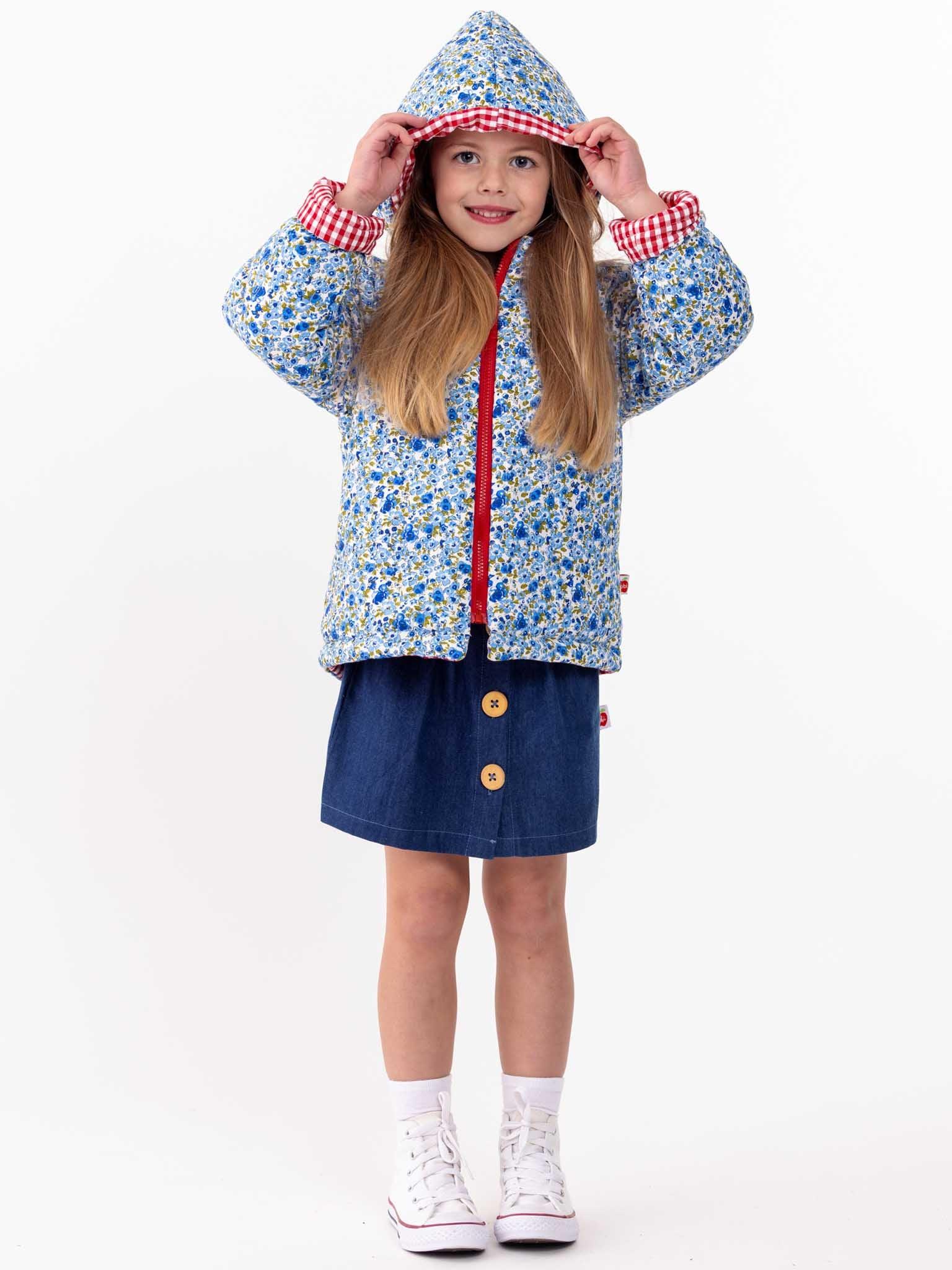 A young girl in an English Wildflowers Floral, Quilted Hooded Reversible Jacket and denim skirt, wearing white sneakers, playfully holding the hood of her jacket. She stands against a plain white background.