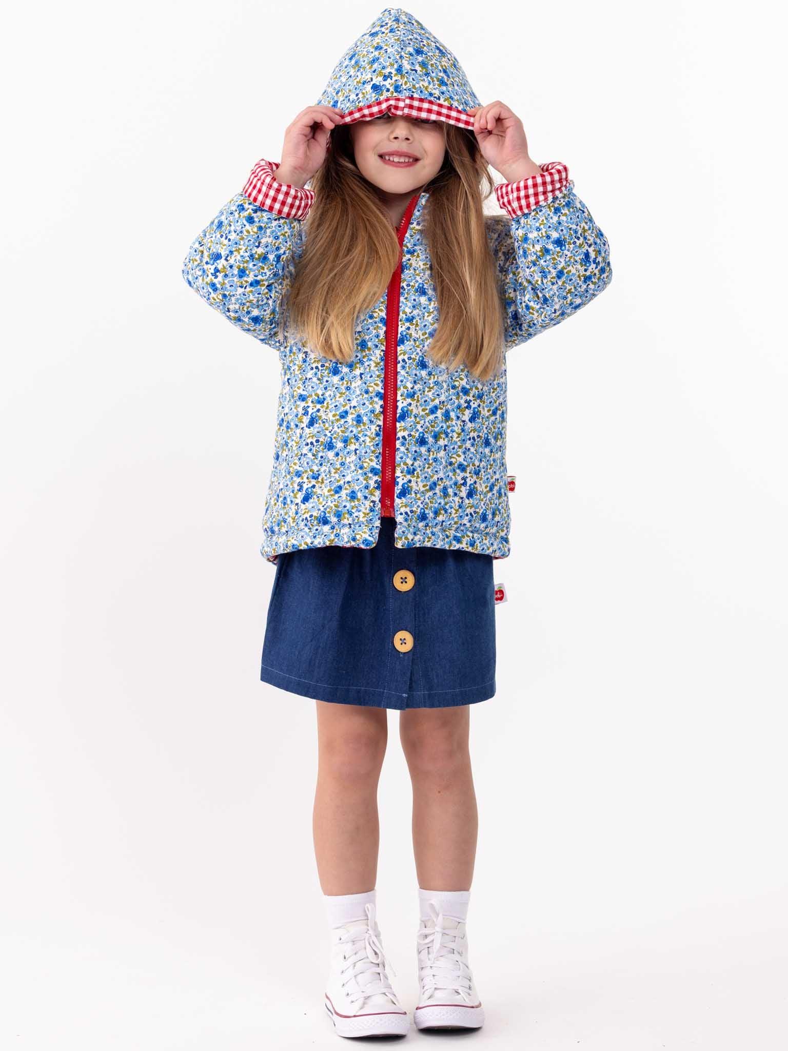 A young girl smiles beneath the hood of her colorful English Wildflowers English Flowers Hooded Reversible Jacket, paired with a denim skirt and white sneakers, against a white background.