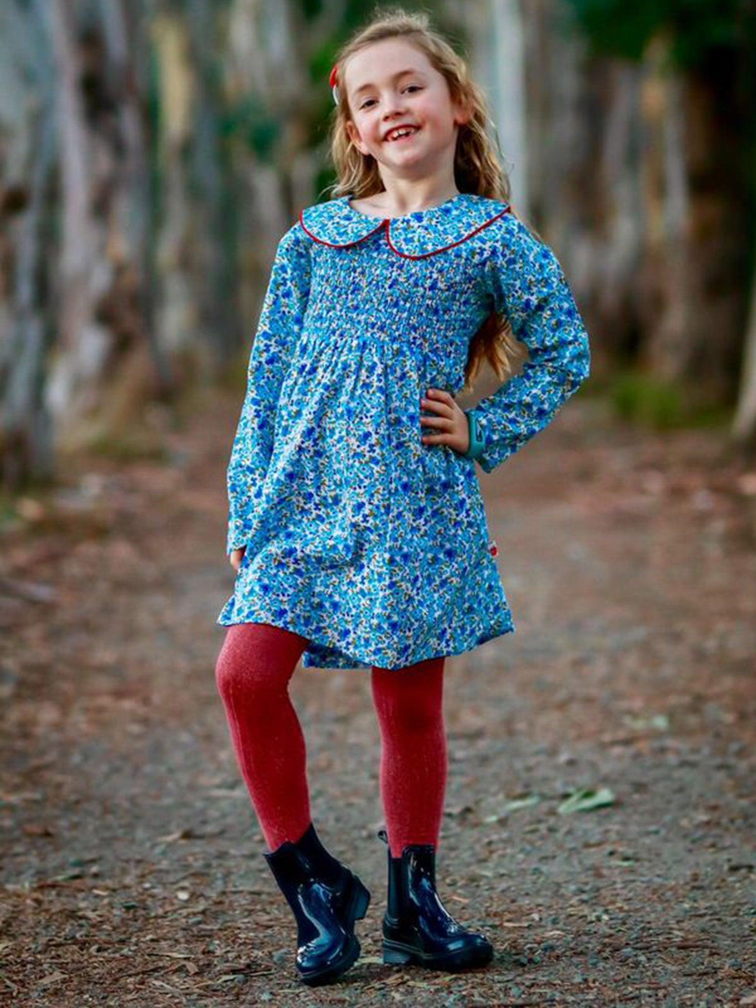 A young girl stands on a forest path wearing the English Flowers Rebecca Dress by English Wildflowers, red tights, and black boots. She is smiling and has one hand on her hip, radiating vintage vibes.