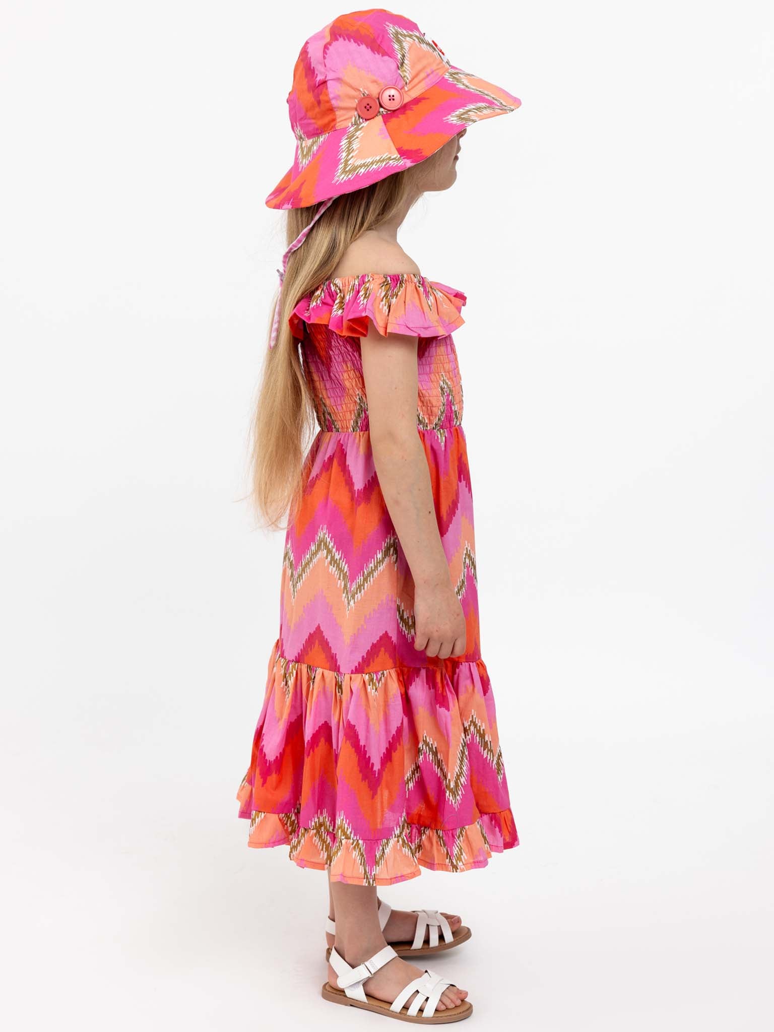 A young girl stands sideways wearing the Golden Ikat Garden Party Dress and a matching hat, both crafted from 100% cotton. She has long blonde hair and is dressed in white sandals, standing against a plain white background.