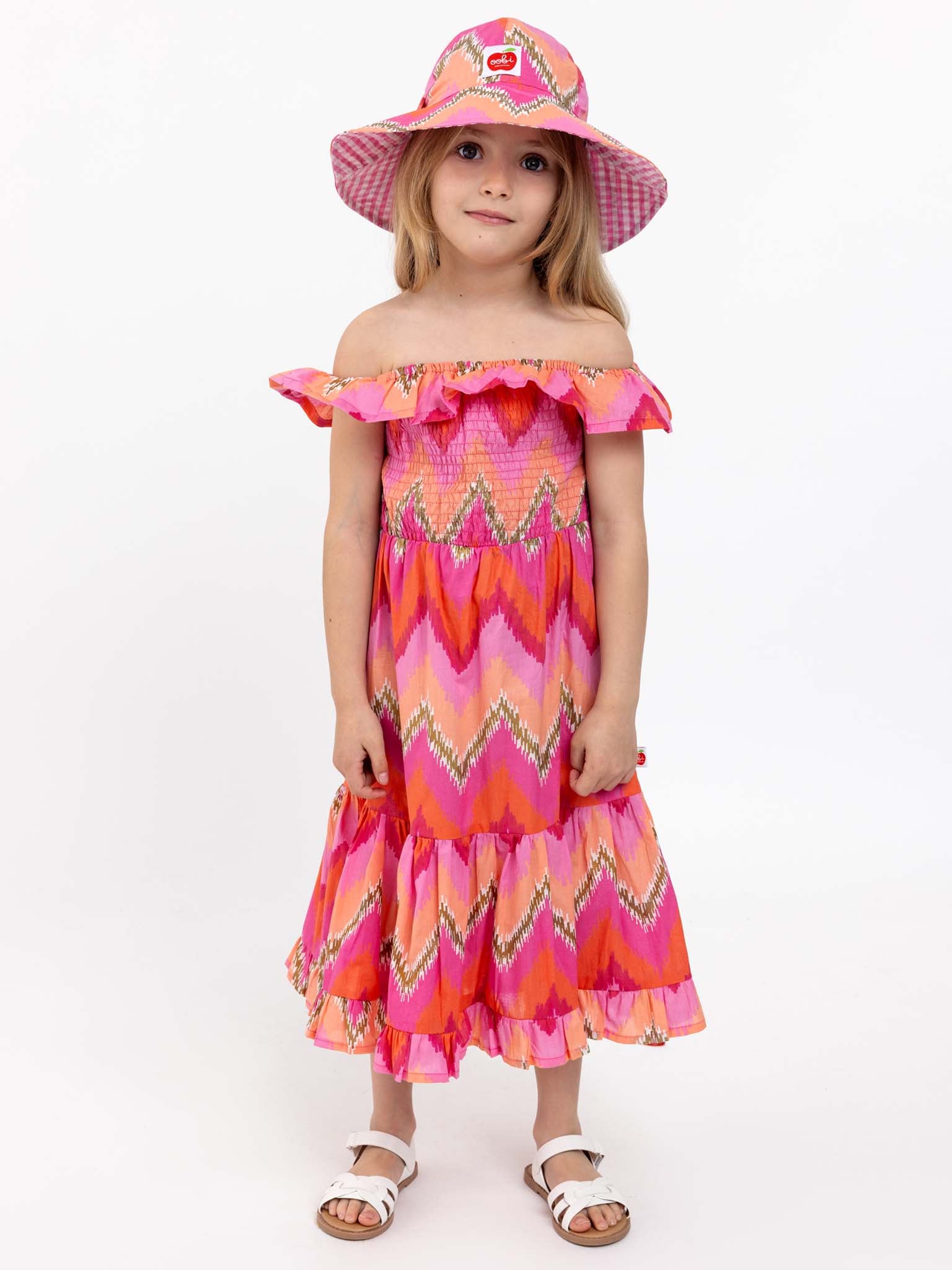 A young girl stands wearing a vibrant Golden Ikat Garden Party Dress featuring a striking zigzag pattern, complemented by a matching wide-brimmed hat and white sandals, set against a plain white background.