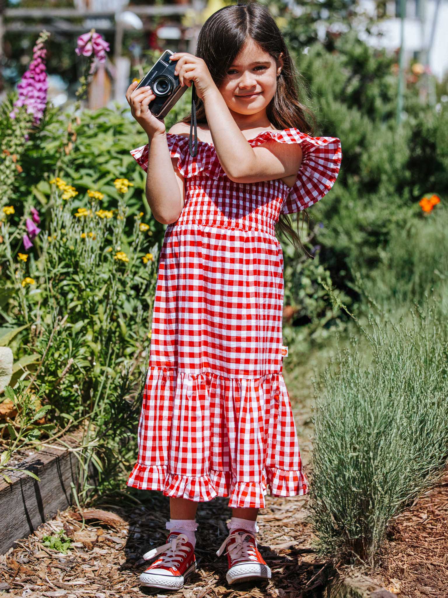 A young girl in a Red Gingham brand Garden Party Dress and red sneakers holds a camera among the flowers and greenery, capturing nature's beauty with every click.