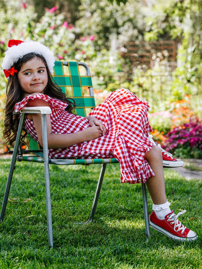 A young girl in a Red Gingham Garden Party Dress and Converse shoes sits on a lawn chair outdoors, smiling with a festive hat. Flowers and greenery bloom vividly in the background, enhancing the charm of her 100% cotton attire from the Red Gingham brand.