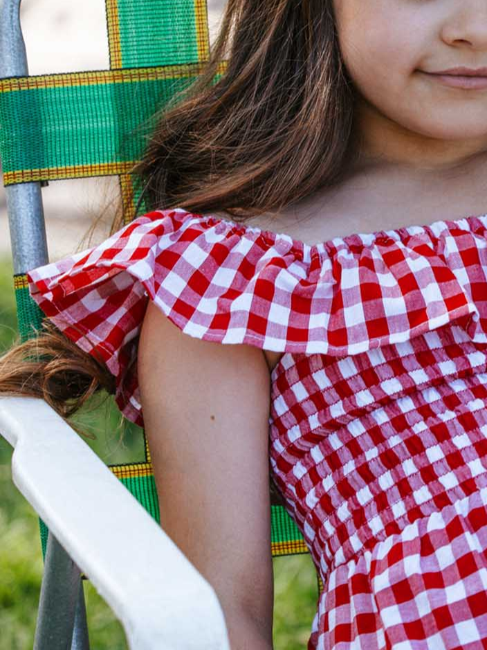 A child in a lively Red Gingham brand dress from their Garden Party collection joyfully sits on a green and yellow lawn chair, embodying the perfect blend of playful charm and comfort.