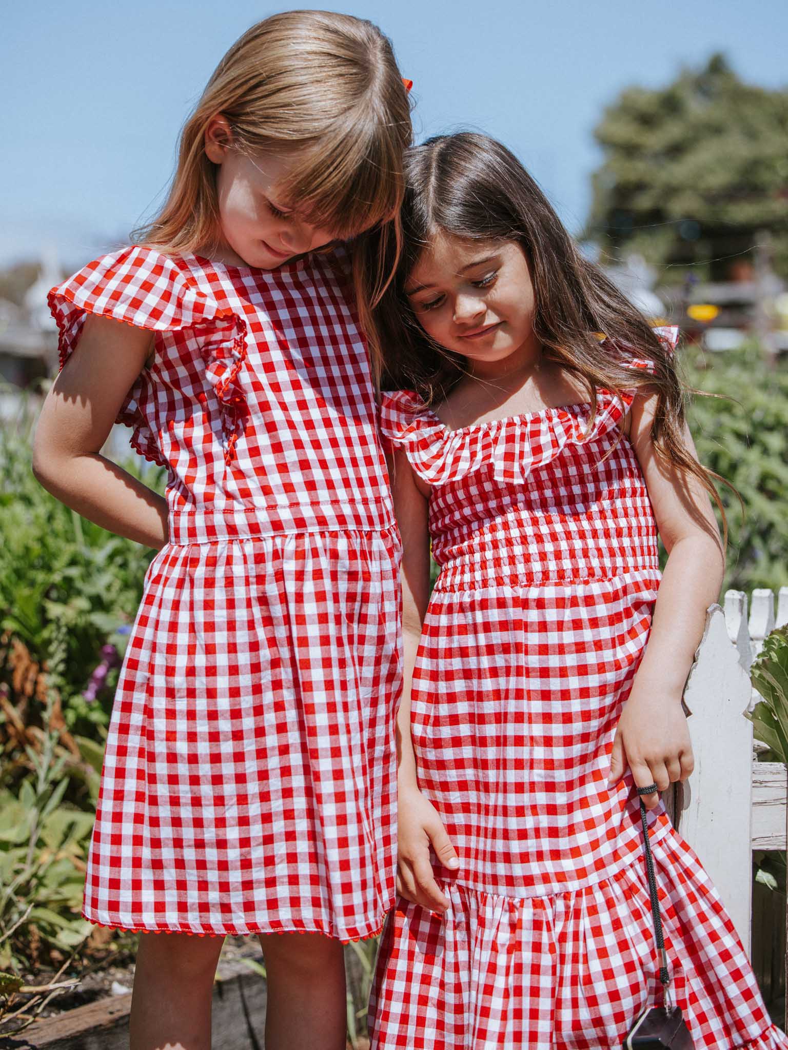 Two young girls stand closely together outdoors, wearing Red Gingham Garden Party Dresses made from 100% cotton.