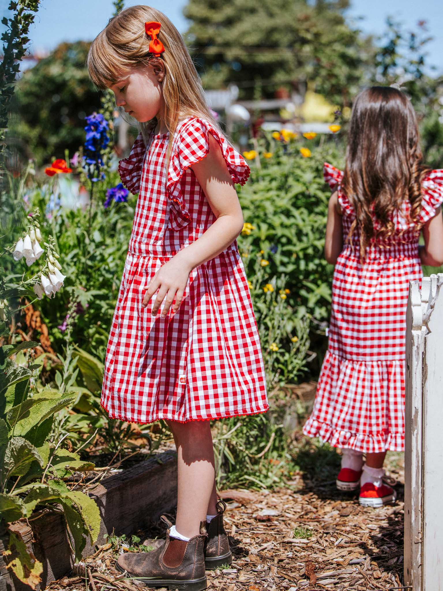 In a vibrant garden, two young girls wearing Red Gingham Lace Edge Dresses with ruffled sleeves are surrounded by blooming flowers. One faces forward while the other turns away, capturing the spirit of carefree summer days under a bright sky.