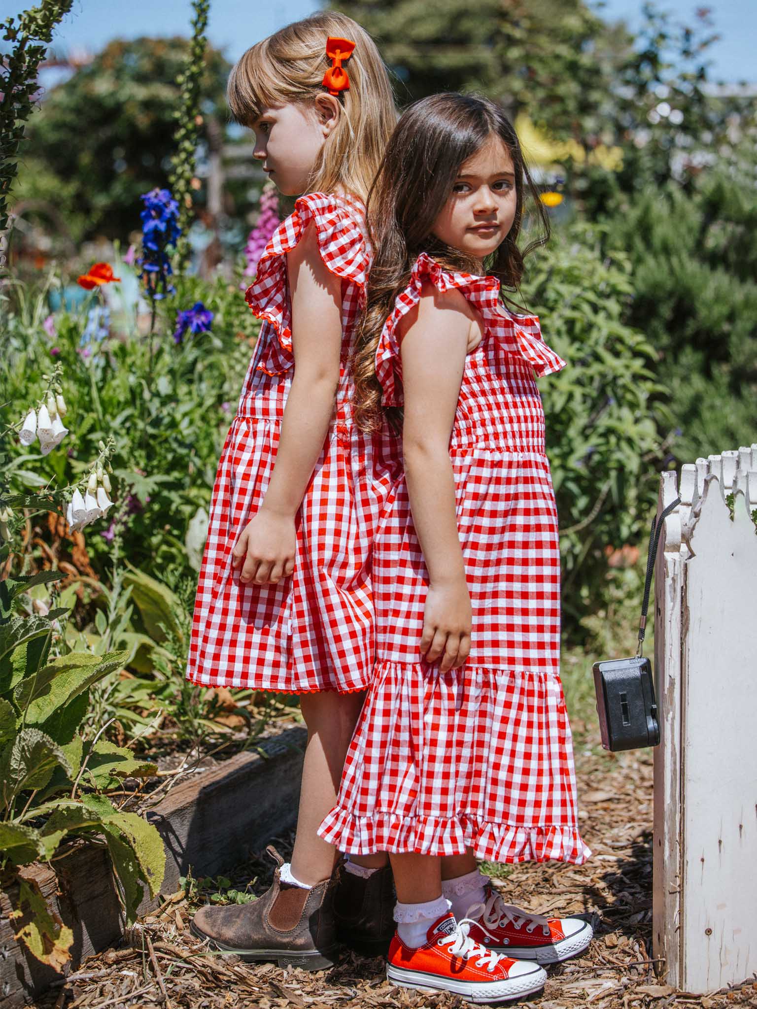 In a lush garden, two young girls wearing Red Gingham's Garden Party Dresses in red stand back to back. One of the dresses is highlighted by a stylish hair clip, while the other outfit is completed with vibrant red sneakers. The 100% cotton fabric effortlessly complements the surrounding greenery and flowers.