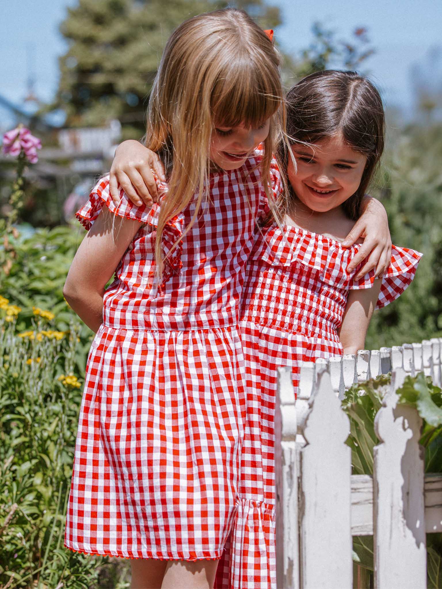 Two young girls in charming garden party dresses from Red Gingham, featuring ruffly sleeves and lace-trimmed hems, stand together in a garden, with one girl's arm around the other.