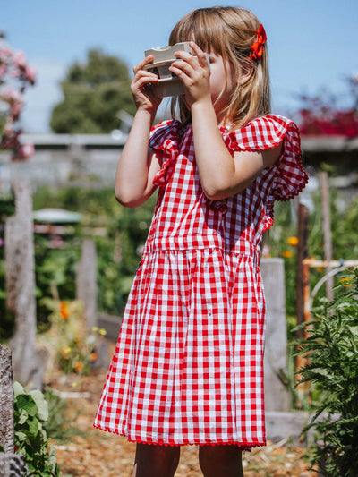 A young girl in a Red Gingham Lace Edge Dress, featuring lace-trimmed, red and white checkered fabric with ruffly sleeves, looks through a viewfinder outdoors, surrounded by plants and flowers.