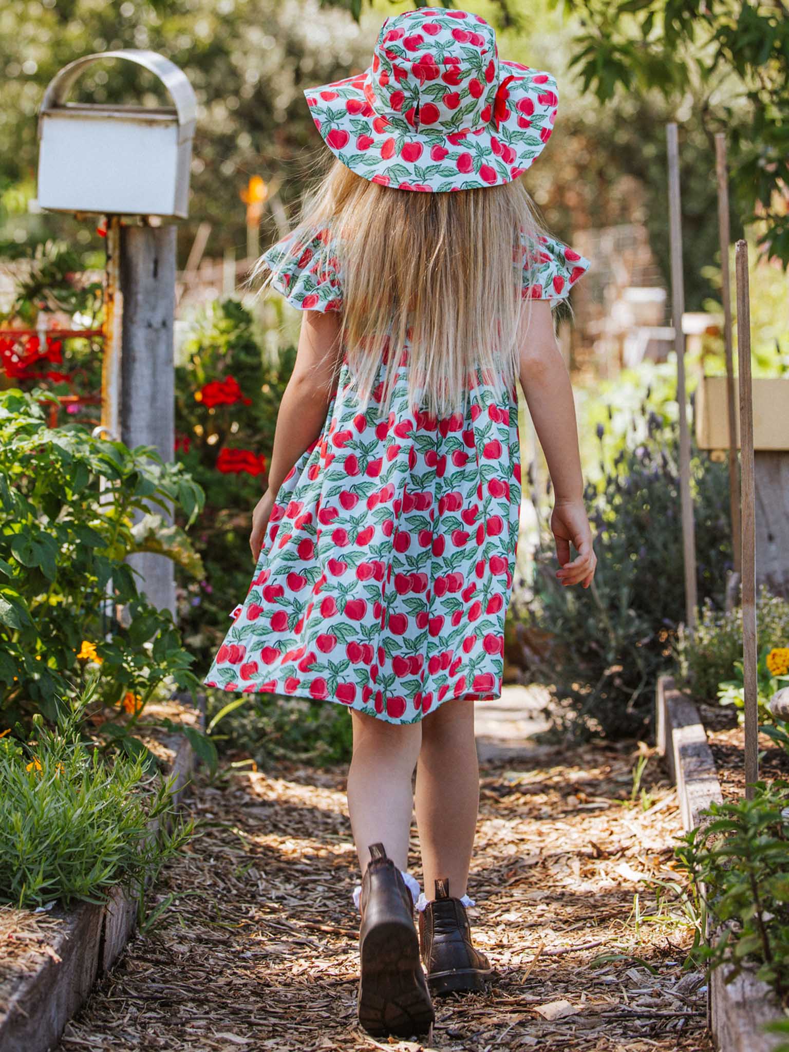 A child wearing a strawberry-patterned dress and a Blue Cherry Chloe Hat strolls down a garden path surrounded by plants.