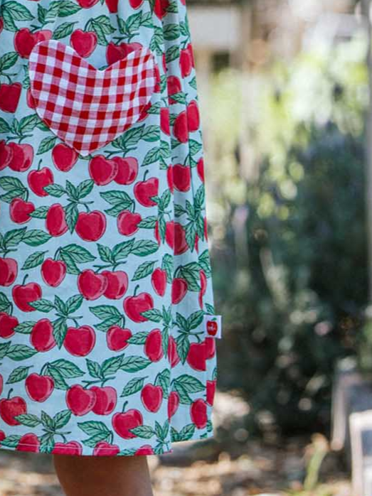 A person stands by the sunny kitchen window, wearing the Blue Cherry Heart Pocket Dress. The vibrant red apple pattern on the apron is beautifully complemented by charming red and white checkered heart-shaped pockets, giving the scene a delightful summer flair.