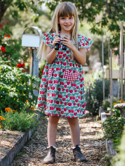 A young girl wearing the Blue Cherry Heart Pocket Dress from Blue Cherry stands on a summer garden path, holding binoculars amidst lush green foliage and vibrant flowers.