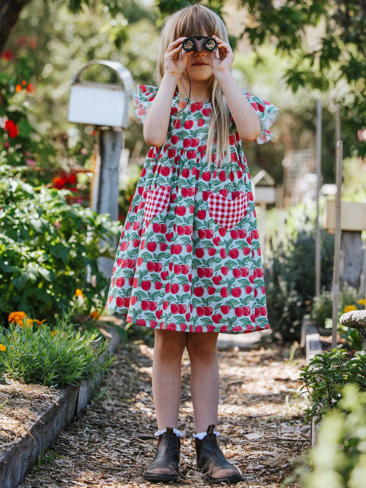 A child dressed in the Blue Cherry Heart Pocket Dress, designed by Blue Cherry, uses binoculars while standing on a sunlit garden path. Surrounded by vibrant plants and flowers, this scene perfectly captures the essence of summer.