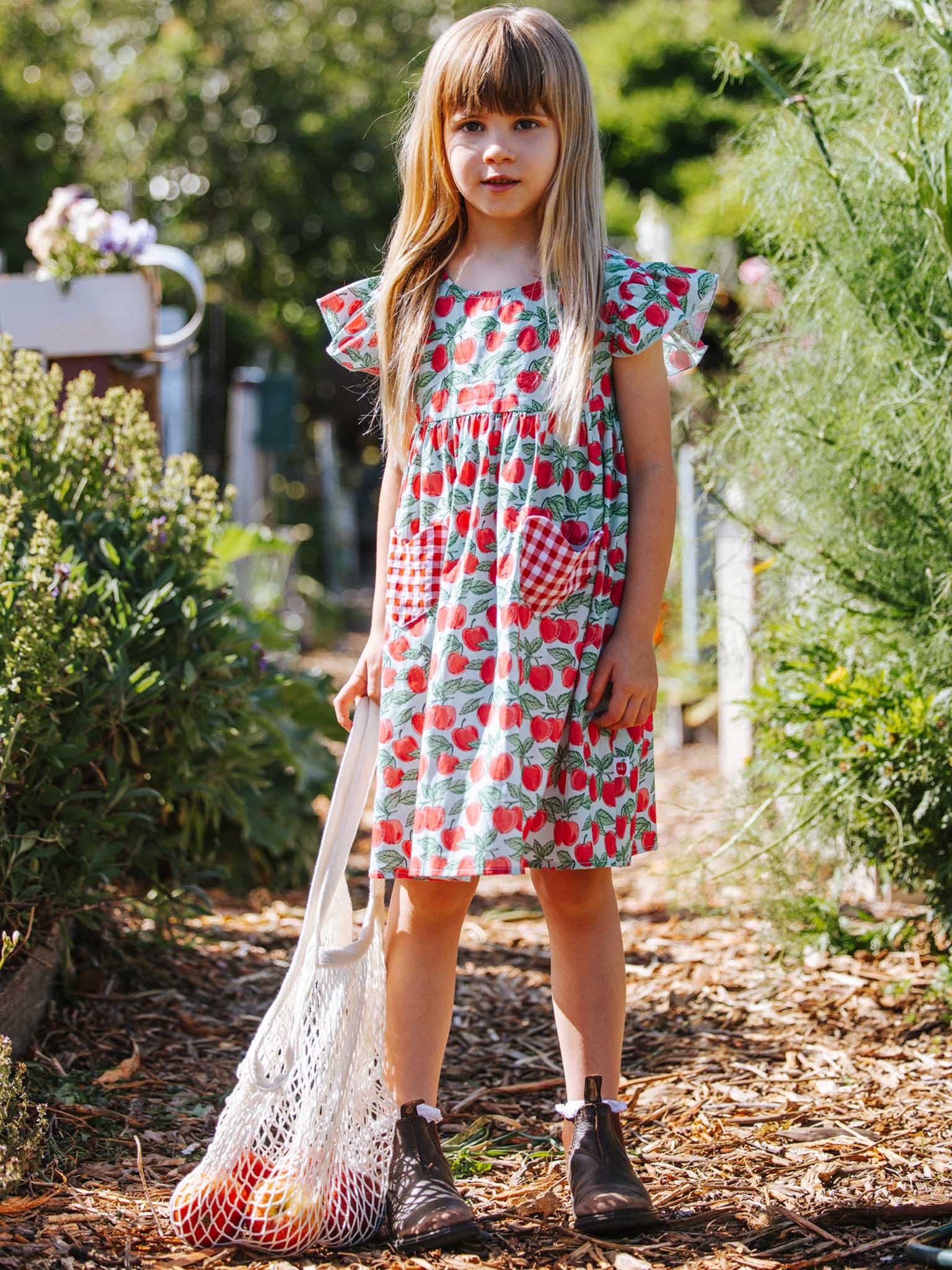 A young girl wearing the Blue Cherry Heart Pocket Dress by Blue Cherry stands on a garden path, holding a mesh bag of apples. Her long blond hair cascades over her brown boots, surrounded by vibrant green plants and an aqua watering can, capturing the essence of summer.