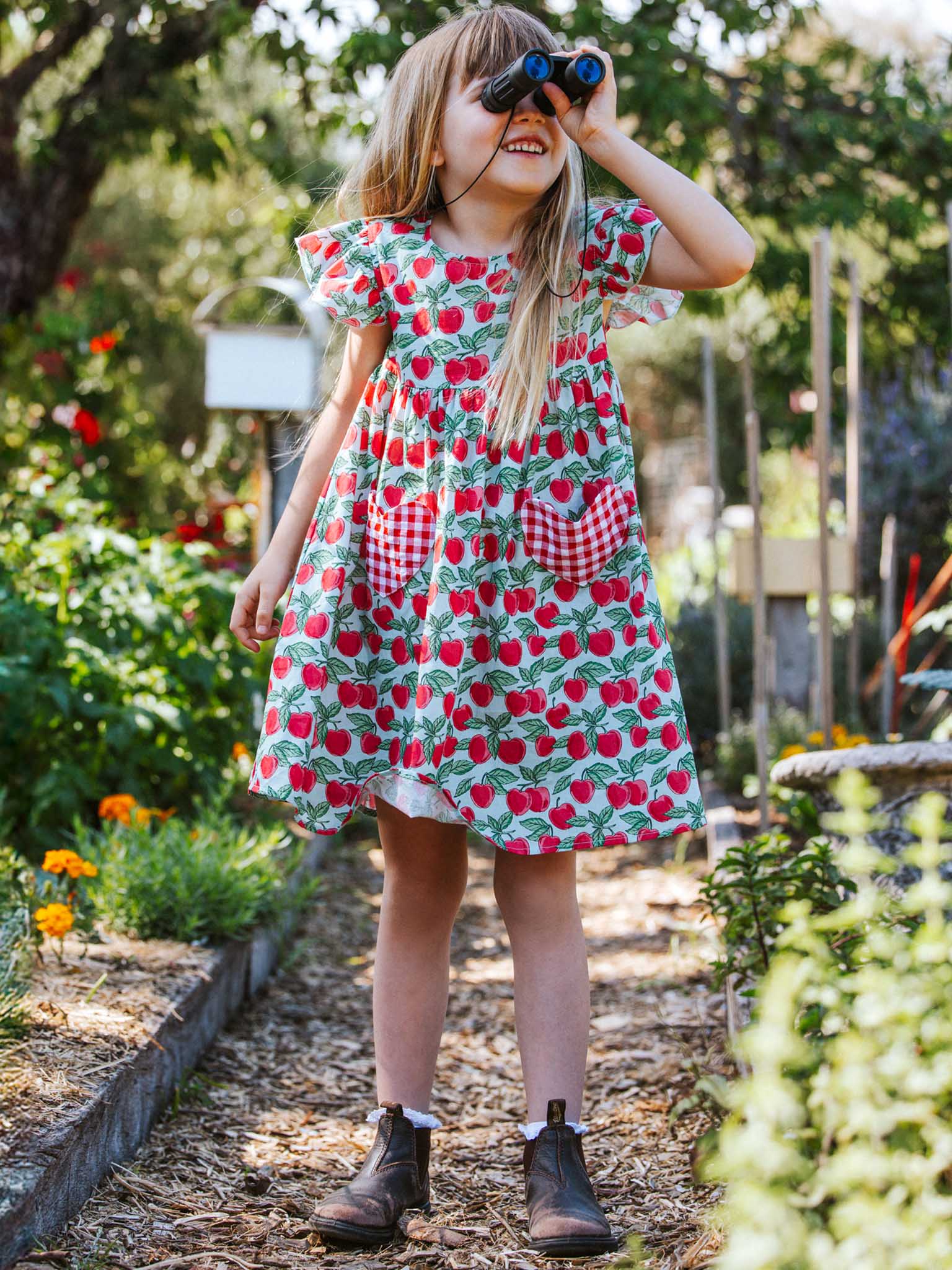 A young girl in a Blue Cherry Heart Pocket Dress paired with boots looks through binoculars in a vibrant summer garden setting.