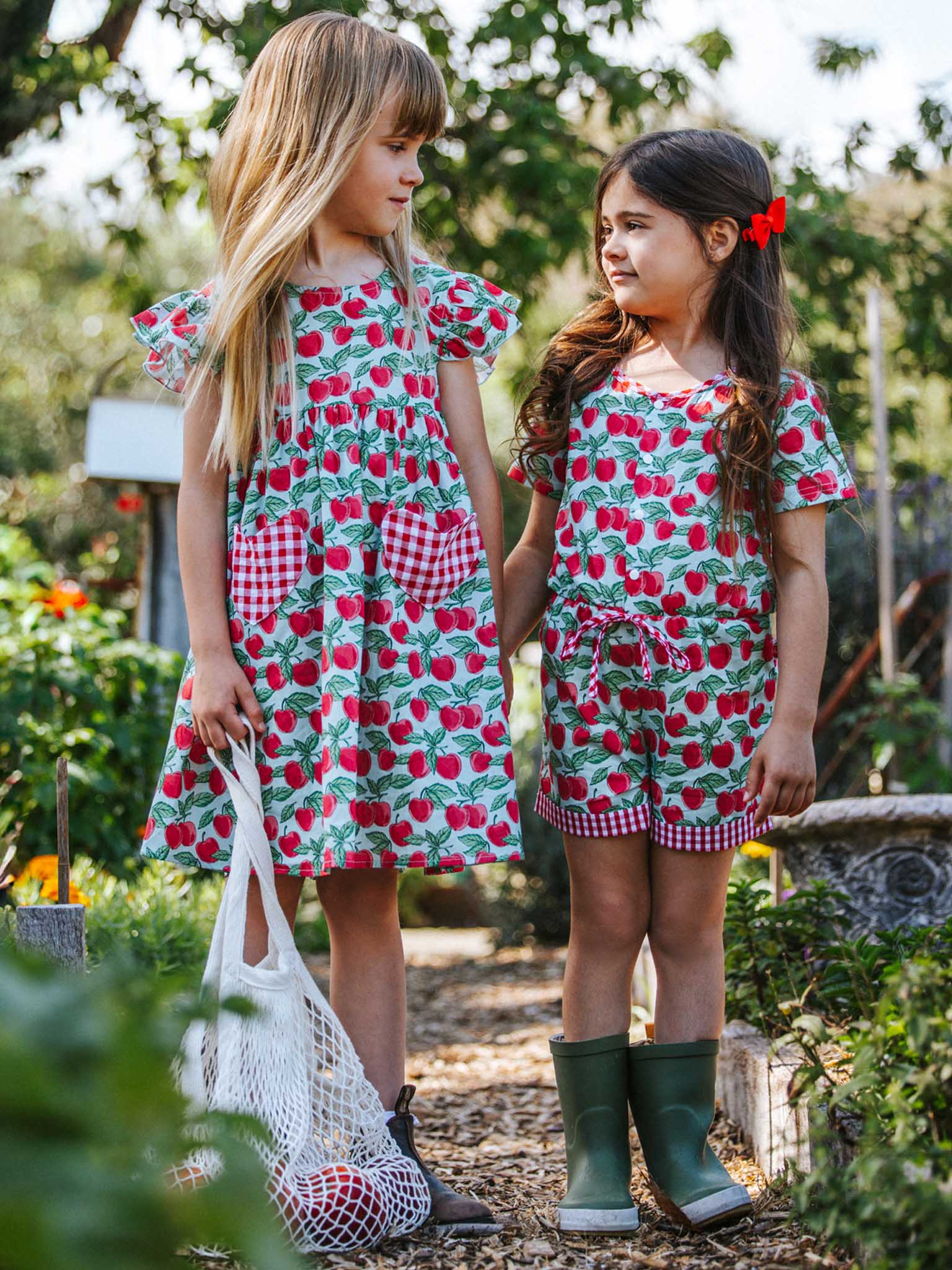 Two girls stand outdoors in matching Blue Cherry Heart Pocket Dresses, featuring playful heart-shaped pockets and vibrant strawberry patterns. In vivid shades of aqua and blue, one girl complements the outfit with boots and a net bag, perfectly capturing the summer spirit.