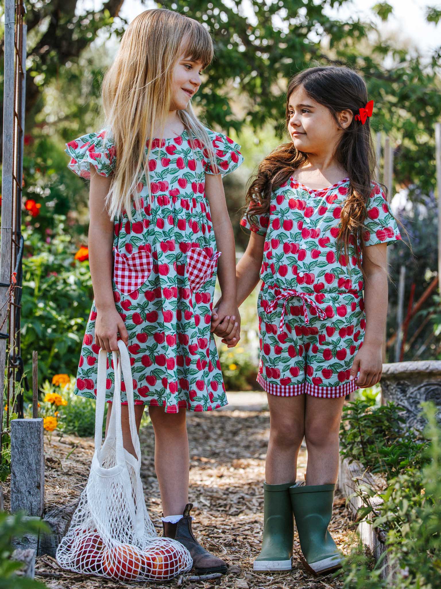 Two young girls in matching Blue Cherry Maisy Playsuits stand outdoors. One holds a mesh bag filled with produce while the other wears green boots. Surrounded by greenery, their charming ensemble adds a touch of whimsy to their delightful day.