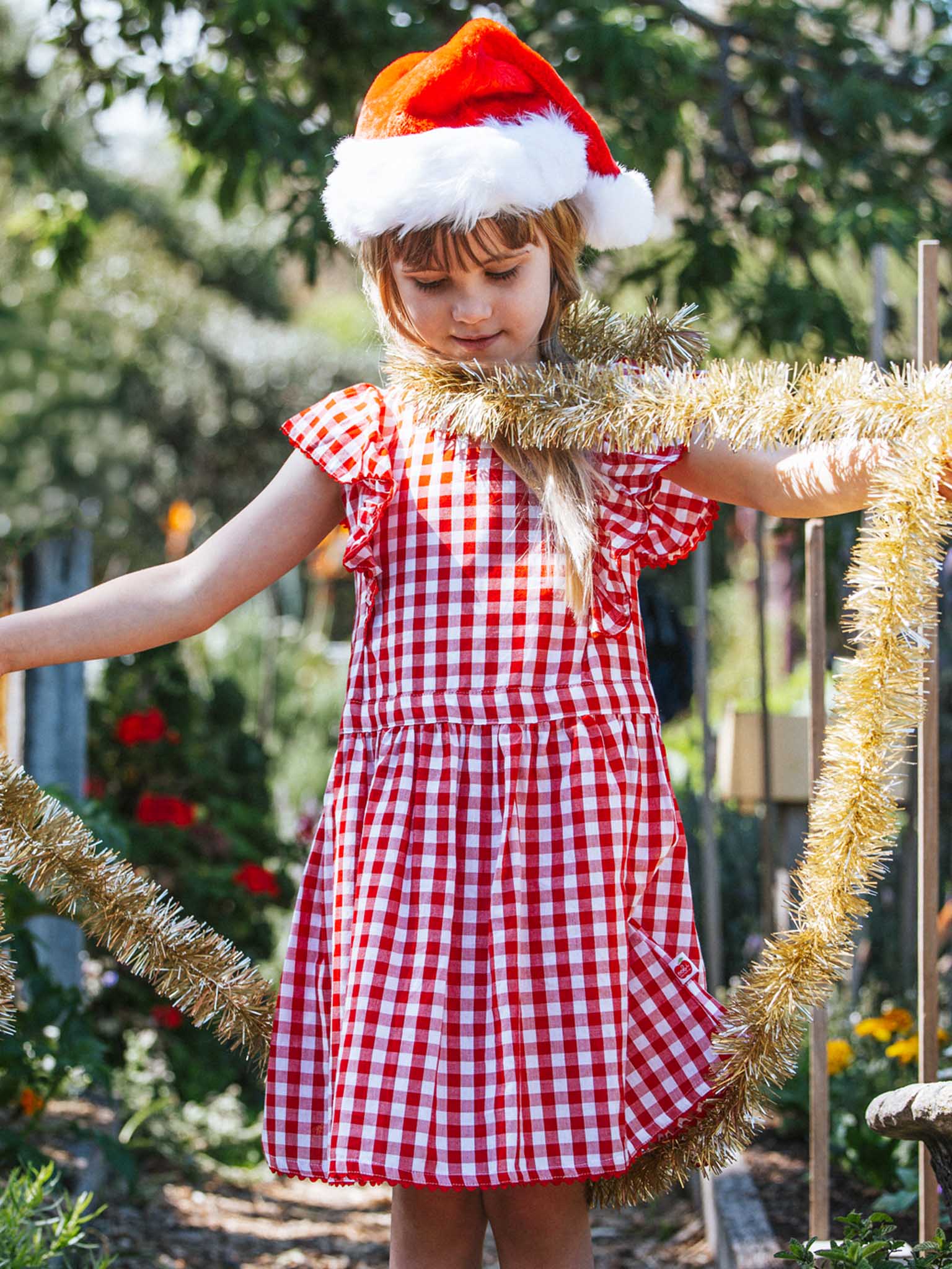 A young girl wearing a Red Gingham Lace Edge Dress with ruffly sleeves and a Santa hat holds gold tinsel, standing outdoors with trees in the background.