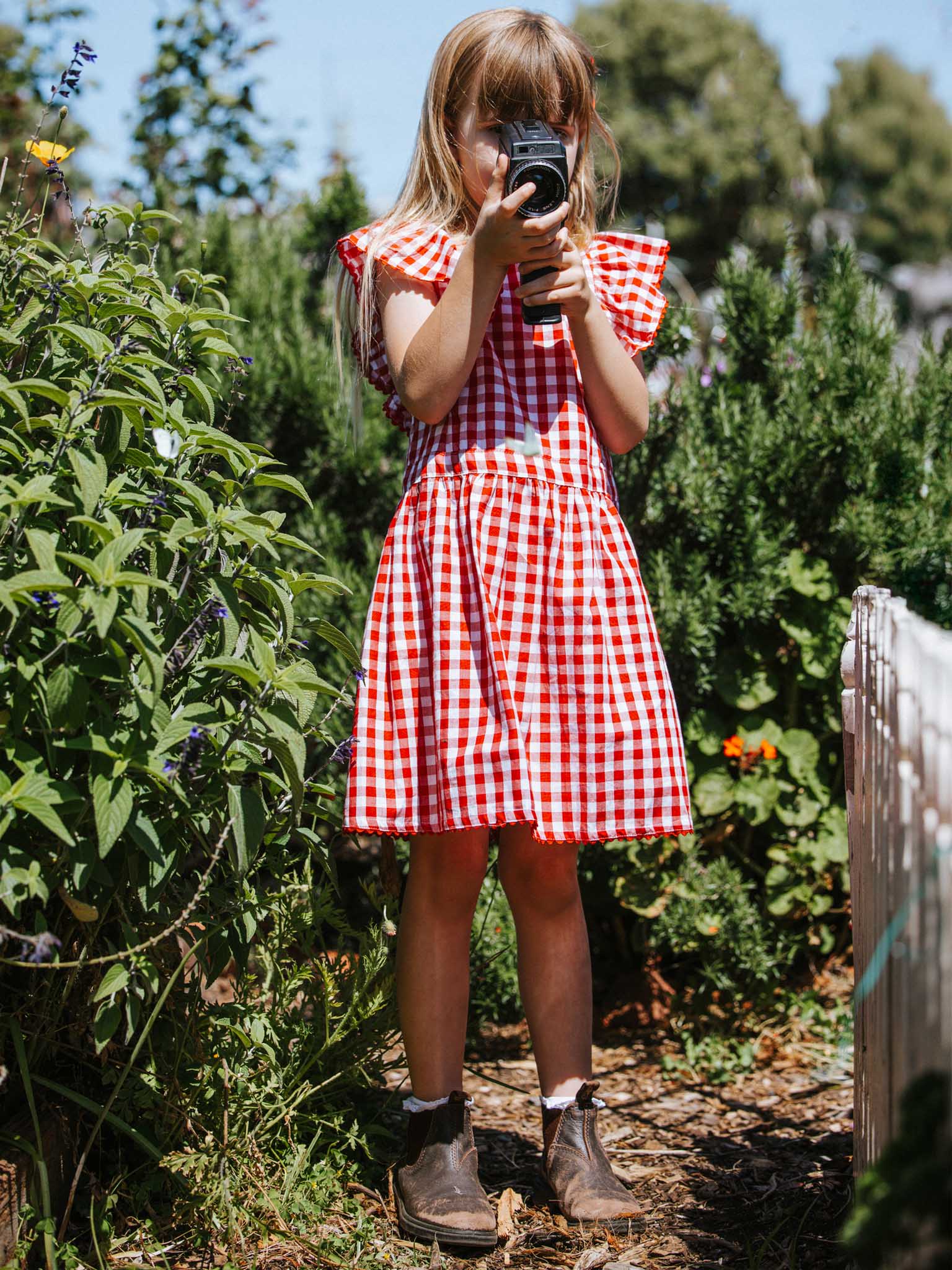 A young girl in a Red Gingham Lace Edge Dress with ruffly sleeves captures a moment with her camera amidst a vibrant garden setting.