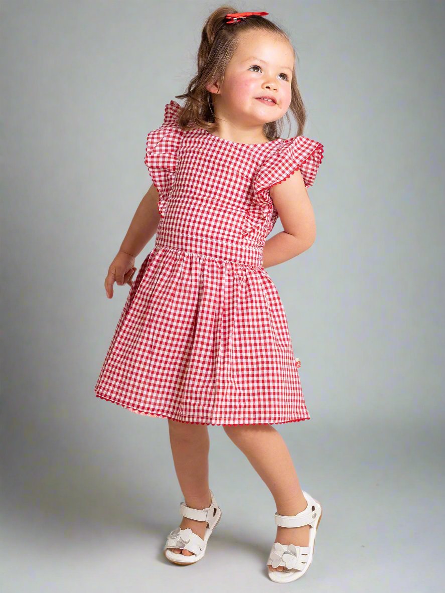 A young girl stands smiling in her Red Gingham Lace Dress & Bloomers, featuring ruffly sleeves and a lace-trimmed hem, with her white sandals completing the charming look against a plain background.