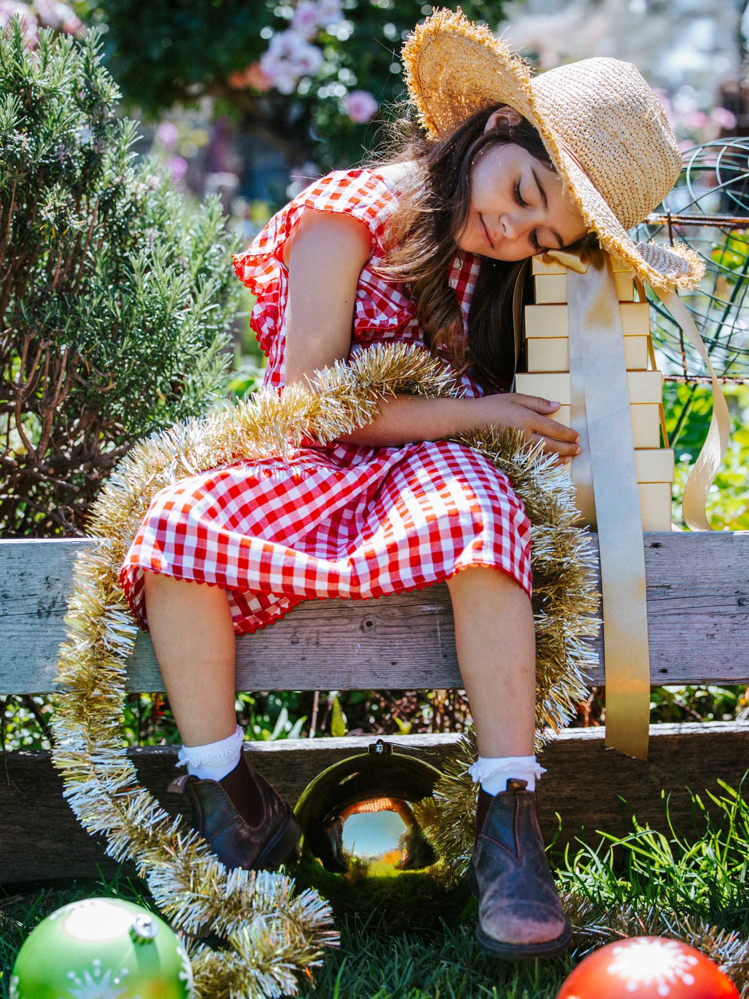 A young girl wearing the Red Gingham Lace Edge Dress paired with a straw hat is holding a stack of ribbon-wrapped presents while seated on a bench decorated with tinsel and Christmas ornaments nearby.