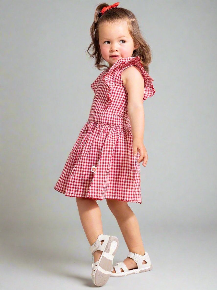 A young girl wearing the Red Gingham Lace Dress & Bloomers, featuring a red and white checkered pattern with ruffled sleeves, poses against a grey background while looking to the side. She complements her outfit with white sandals and a red bow in her hair.