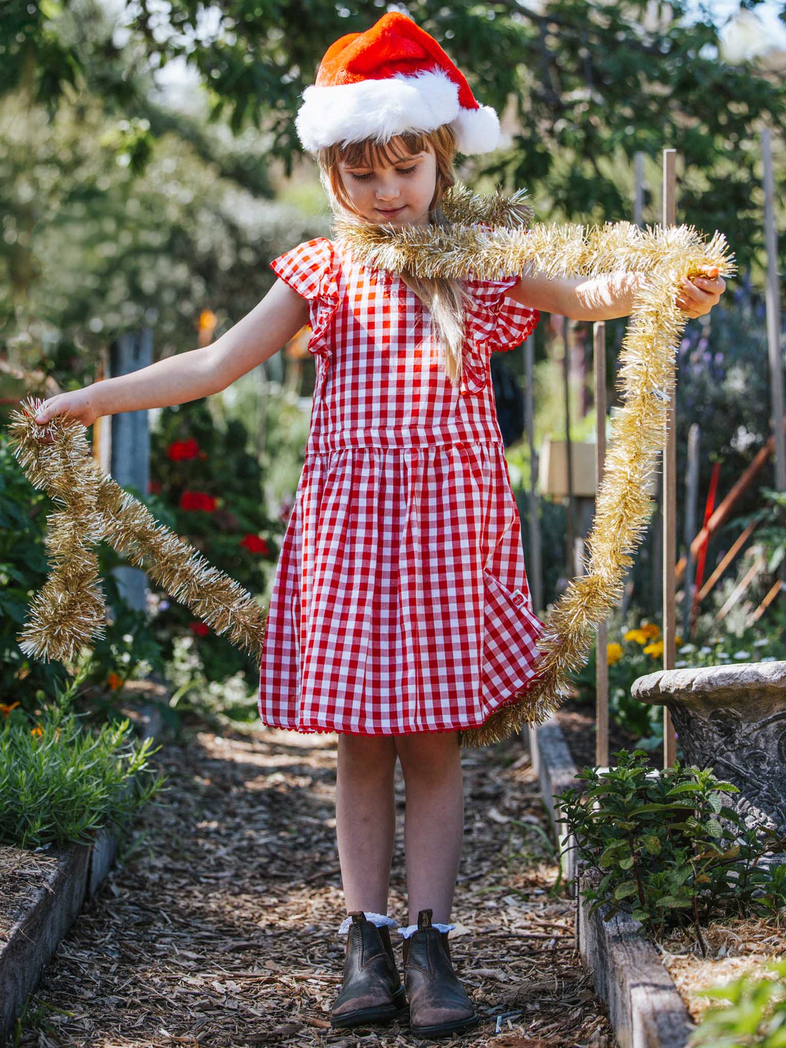 A girl wearing the Red Gingham Lace Edge Dress, featuring lace-trimmed, ruffly sleeves, and a Santa hat is standing in a garden while holding a gold tinsel garland.