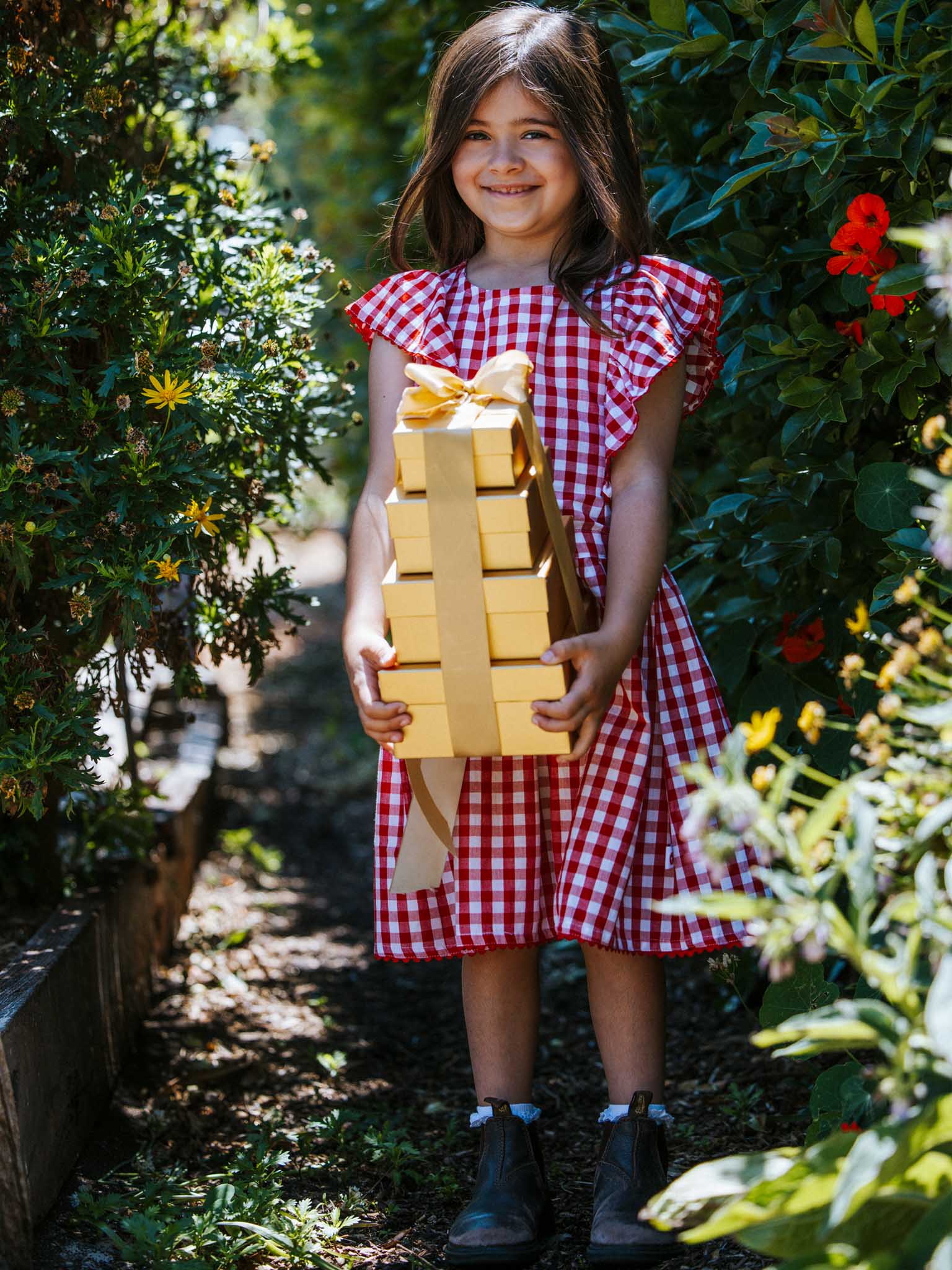 A girl in a Red Gingham lace-trimmed dress with ruffly sleeves stands outdoors, holding a stack of gold-wrapped gifts.