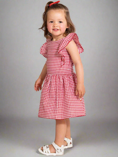 A young child dressed in a captivating Red Gingham Lace Dress & Bloomers stands against a neutral background. The outfit is perfectly paired with white sandals and a coordinating red hair bow.