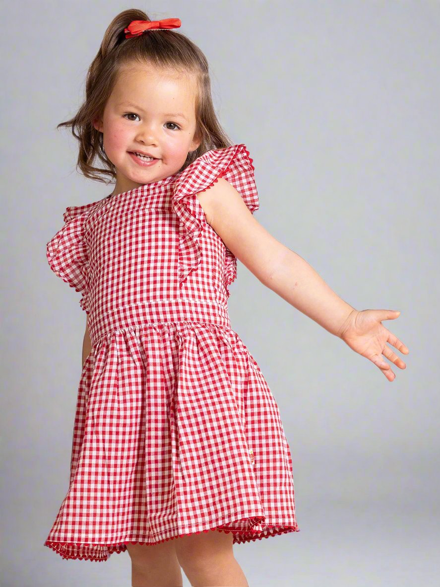 A cheerful child wearing a Red Gingham Lace Dress & Bloomers with ruffled sleeves poses gracefully, arm outstretched, against a simple backdrop.