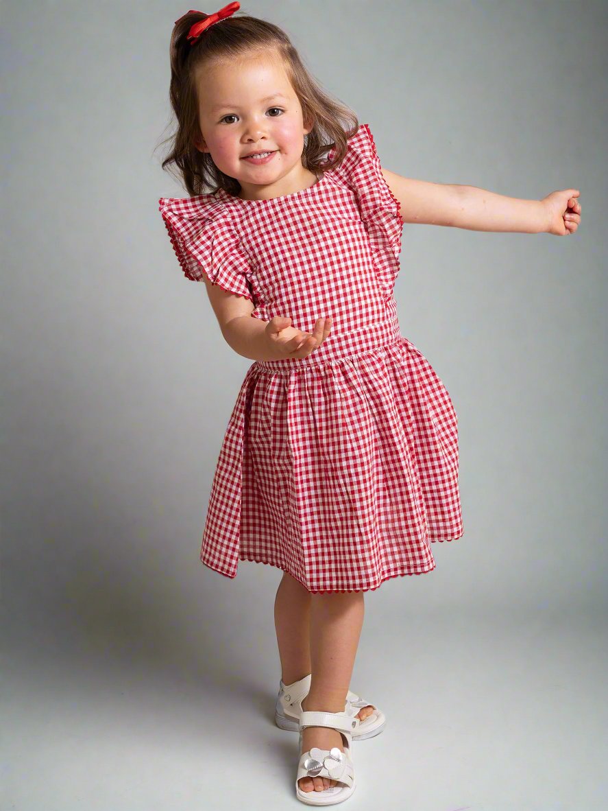 A young girl joyfully poses in a Red Gingham Lace Dress & Bloomers, featuring a playful checkered pattern and ruffled sleeves. She smiles brightly with one arm extended, wearing white sandals and a red bow in her hair.