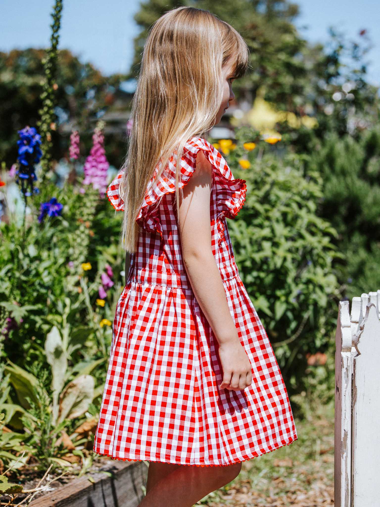 A young girl with long blonde hair, wearing a Red Gingham Lace Edge Dress from Red Gingham, stands in a garden surrounded by colorful flowers.