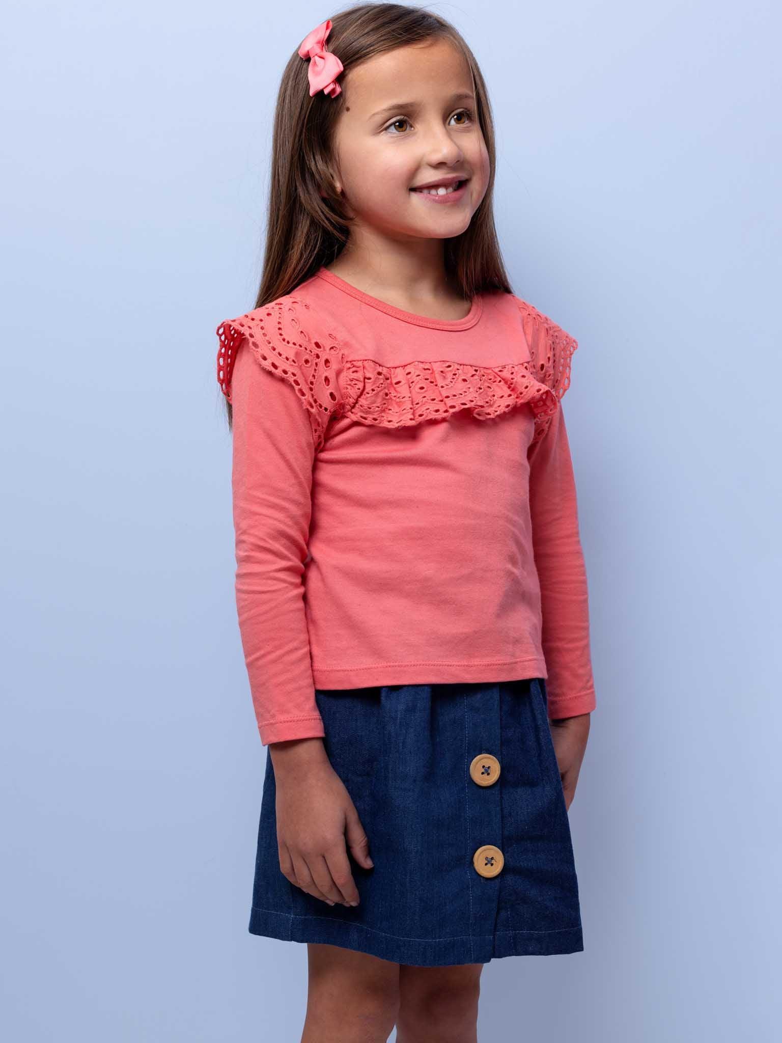 Young girl in an Essentials Lace Front Tee Warm Pink and denim skirt smiling against a light blue background.