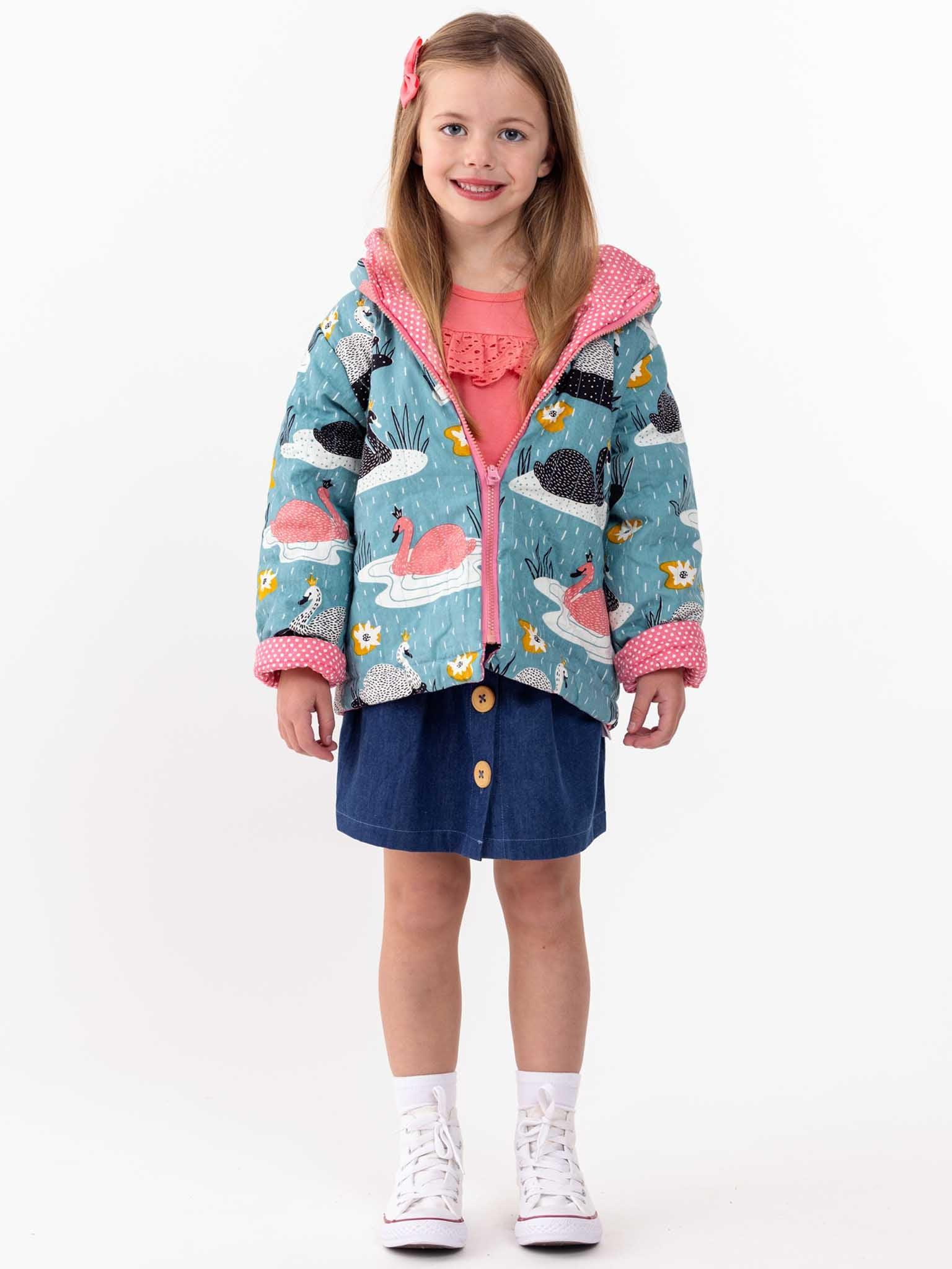 A young girl standing and smiling, wearing a teal jacket with flamingo patterns, a Imperfect lace Front Tee Warm Pink, and a denim skirt against a white background.