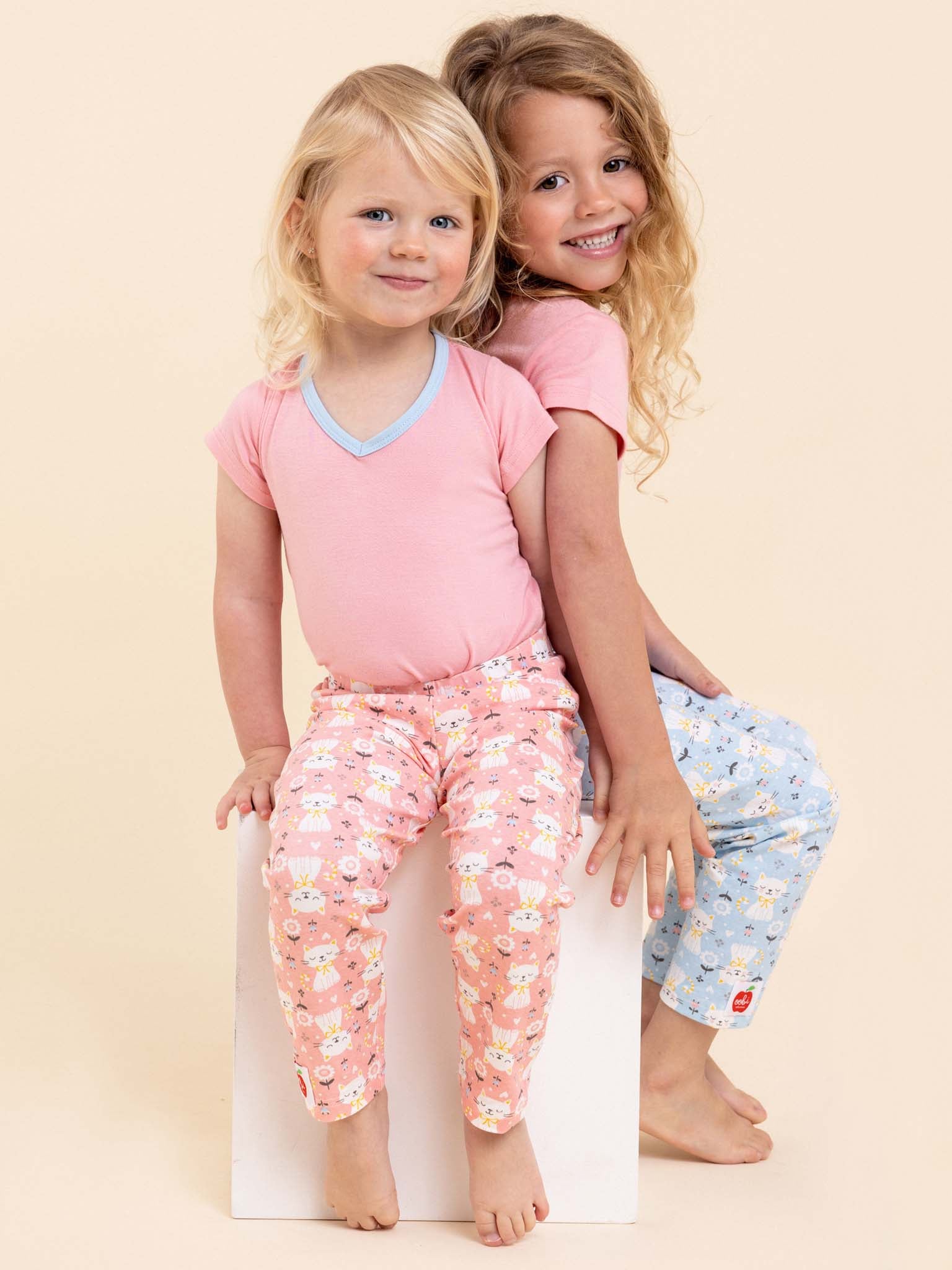 Two young children are sitting on a white cube, wearing the Pink Kitten Lounge Pants & Free Tee by Cat, smiling happily.