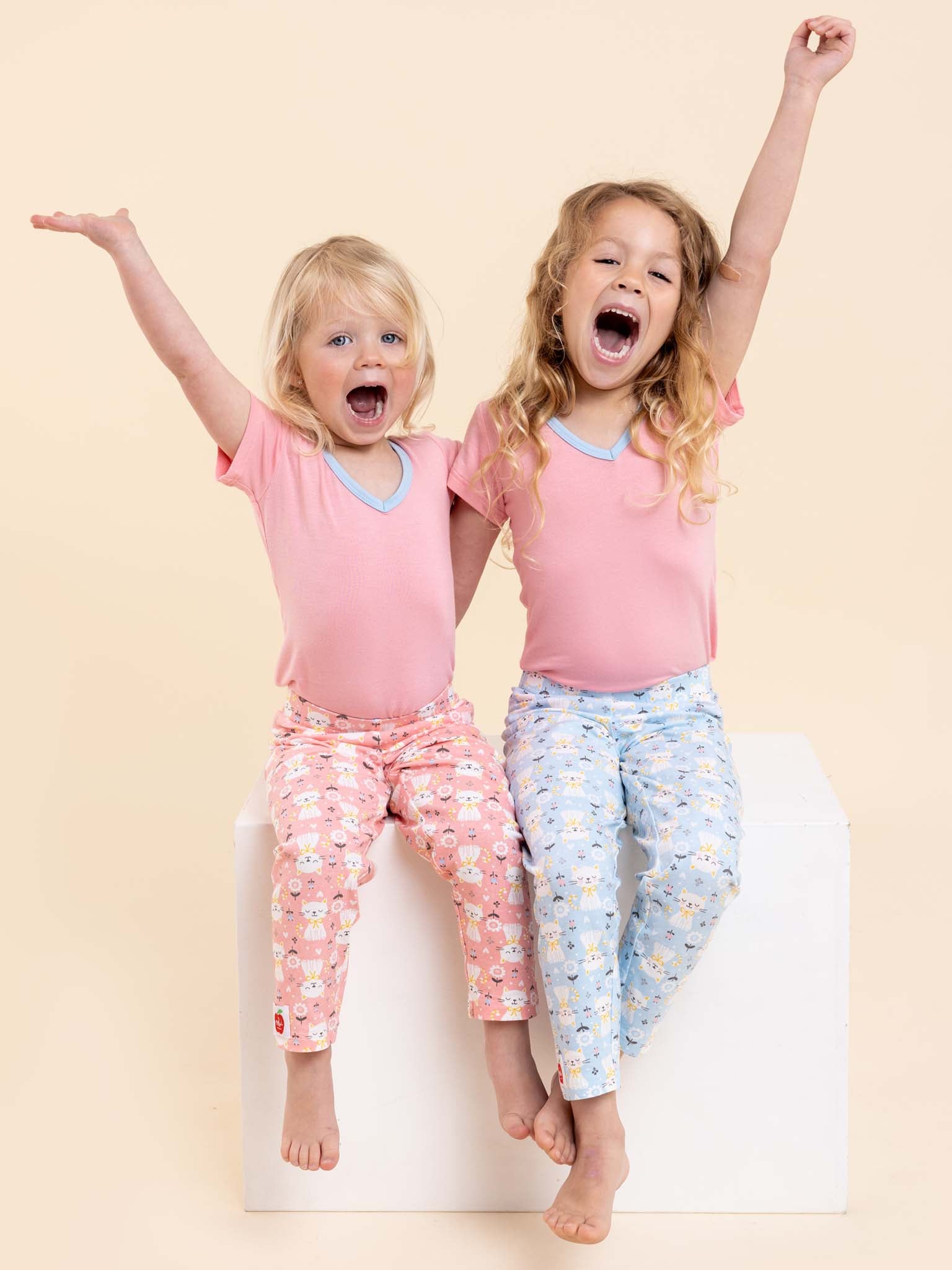 Two cheerful children wearing Cat's Pink Kitten Lounge Pants & Free Tee sit on a white block with arms raised, smiling against a beige backdrop.