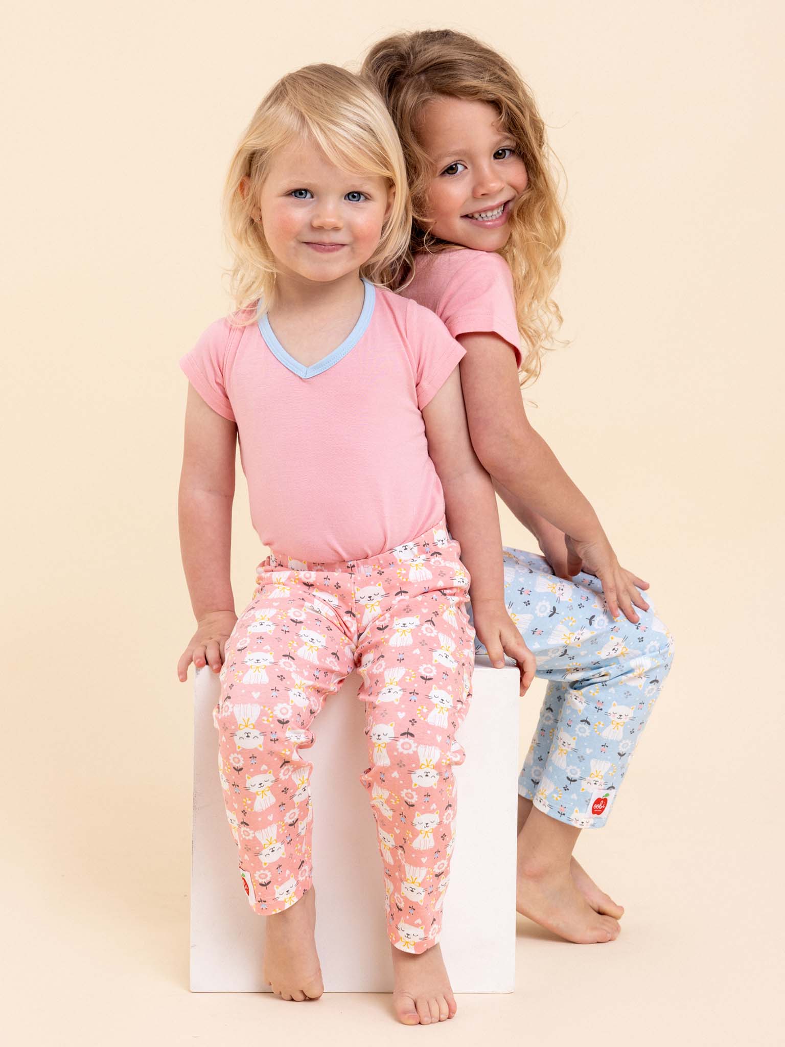 Two young children sit on a white block, smiling in Pink Kitten Lounge Pants and matching free tees by Cat. One wears a light pink design, the other light blue—both embodying the joyful spirit of an Aussie Child donation.