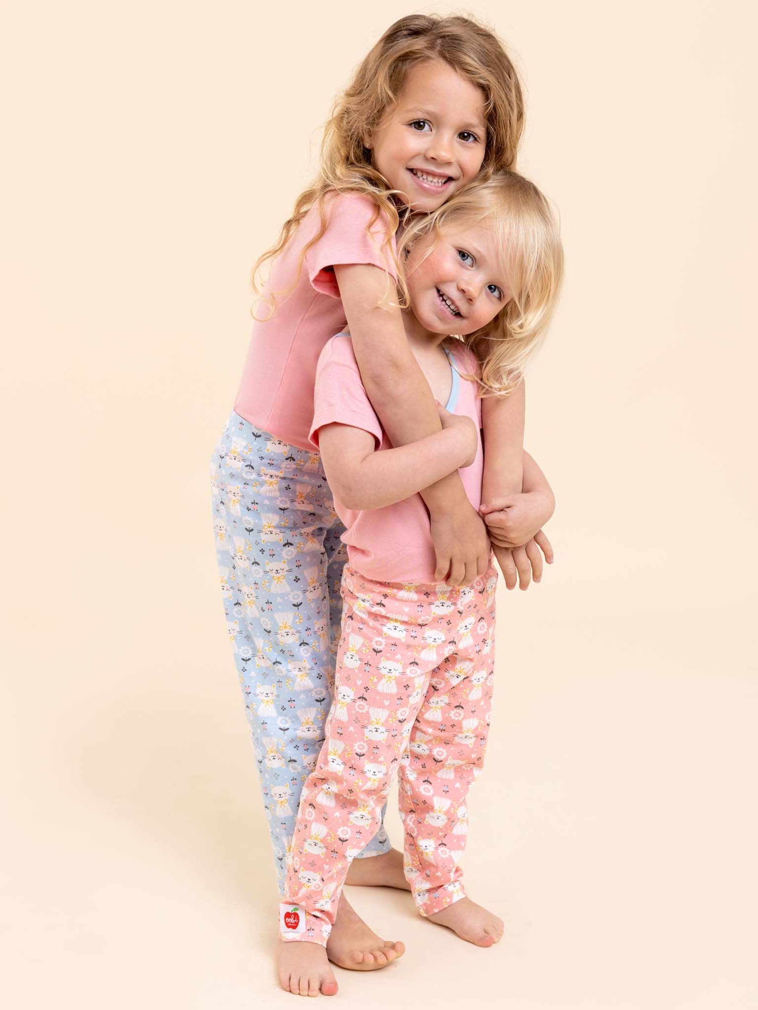 Two young children stand smiling in pink tops and patterned pants. The child in front is hugged from behind. One wears blue cotton jersey leggings, while the other is dressed in Cat's Pink Kitten Lounge Pants & Free Tee—the softest, stretchiest lounge pants available.