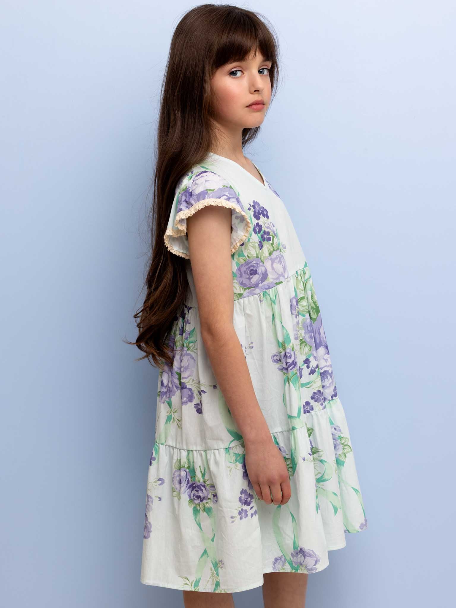 A young girl with long brown hair, wearing an Ocean Lyric Dress adorned with purple and green floral patterns and vintage lace trim, stands against a plain light blue background.