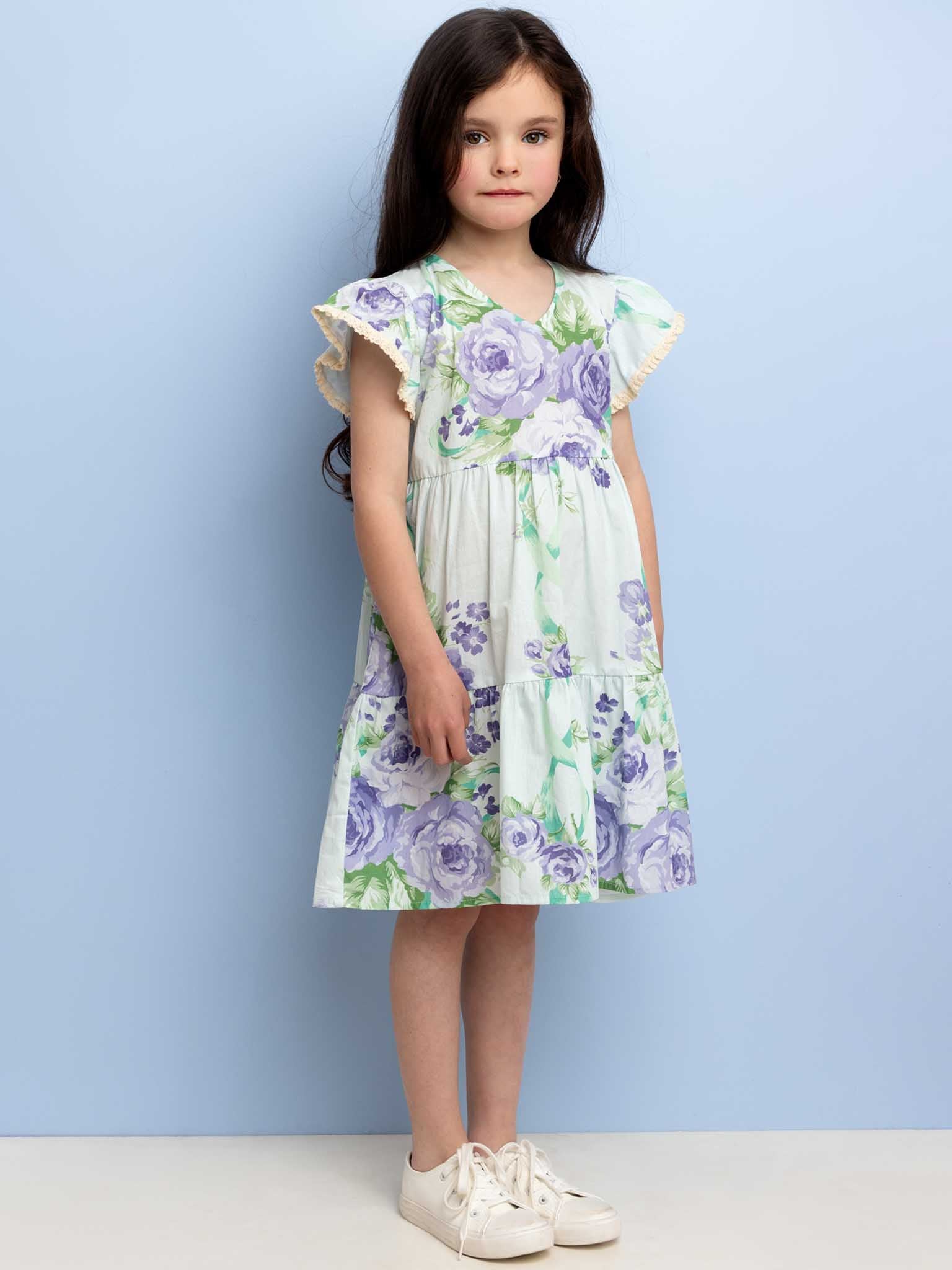 A young girl with long dark hair in an Ocean Lyric Dress adorned with purple floral patterns, standing against a light blue background, and wearing white sneakers.