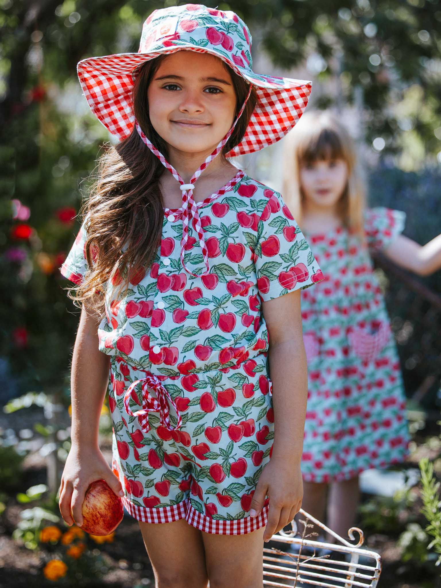 Two girls in a garden, one in the foreground wearing a Blue Cherry Chloe hat featuring a blue cherry pattern, wide brim, and ponytail hole, holding an apple while blurred flowers and foliage create a soft backdrop.