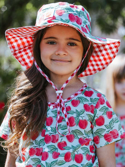 A child beams with joy, sporting a Blue Cherry Chloe hat featuring a wide brim and ponytail hole, perfectly complementing her dress adorned with a playful red apple pattern.