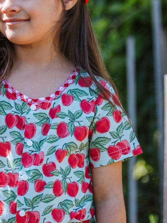 A girl named Maisy stands outdoors, proudly wearing the playful Blue Cherry Maisy Playsuit adorned with a delightful red apple pattern. Her choice of outfit reflects a love for vibrant and fun designs.