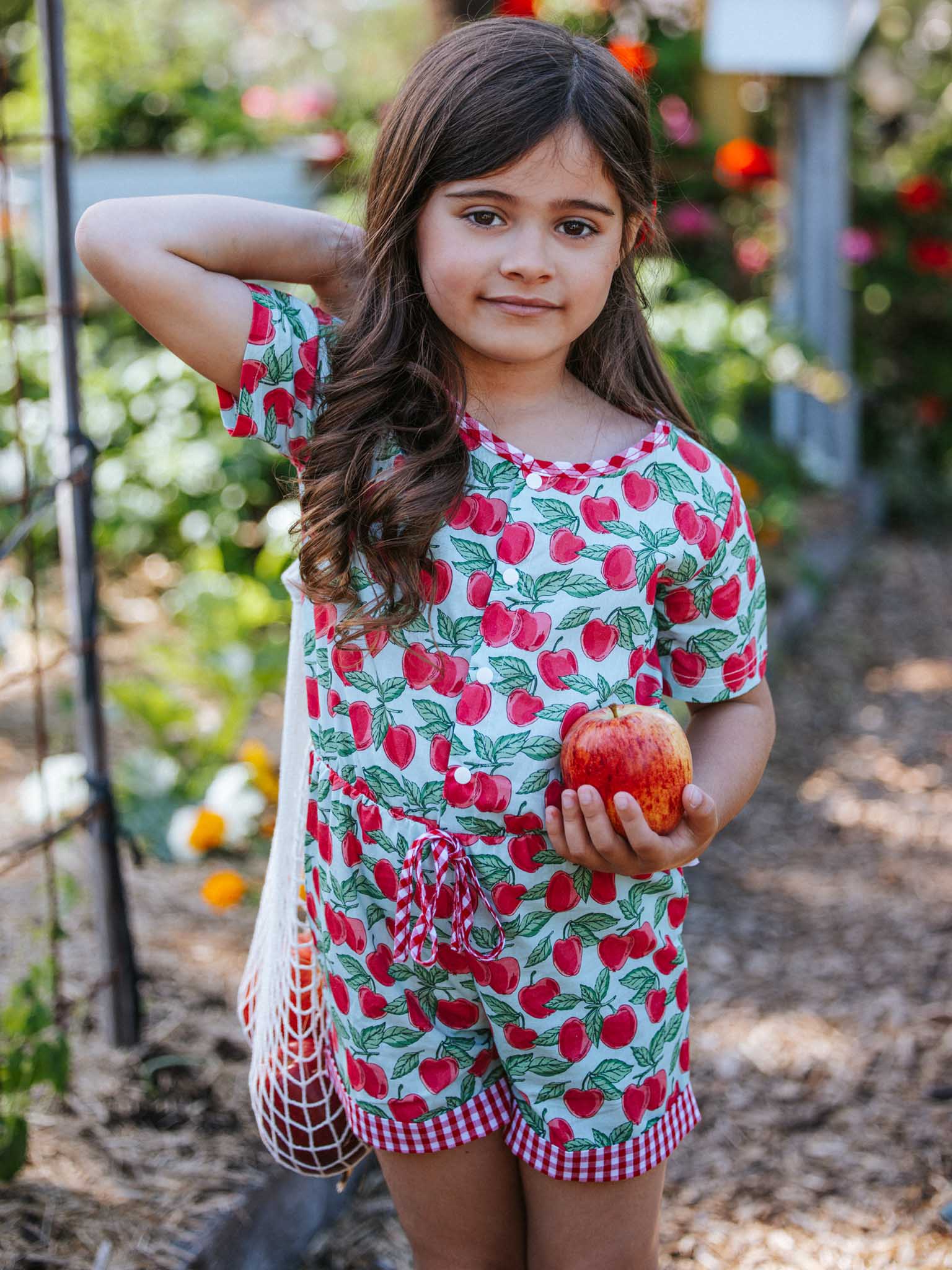 A girl in Blue Cherry's Maisy playsuit holds an apple in one hand while standing in a garden, with a net bag hanging over her shoulder. Her cheerful smile perfectly captures the youthful charm of Blue Cherry's girls' clothes collection.